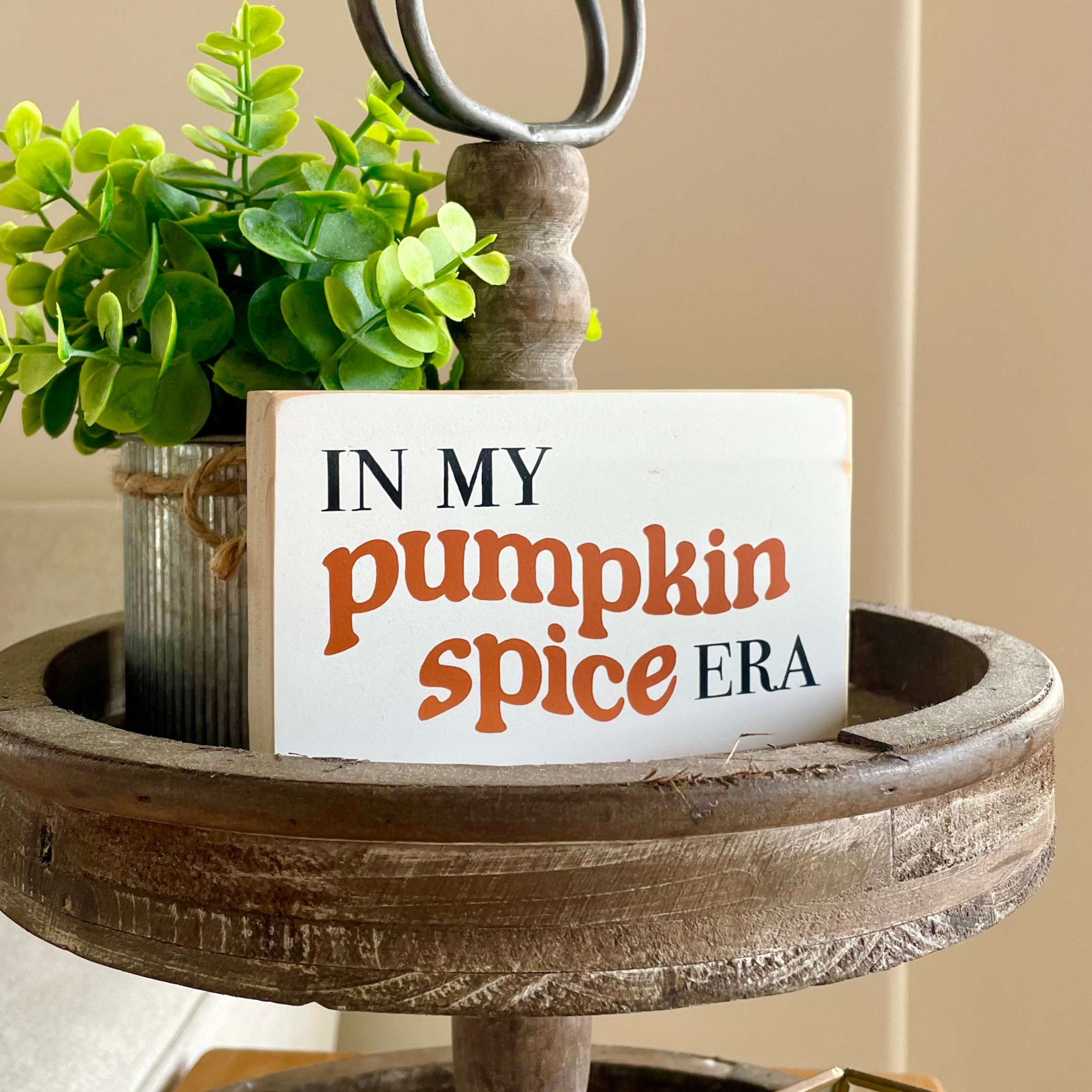 a small, white, wood sign that reads" in my pumpkin spice era" sits on a tiered tray with a plant next to it.