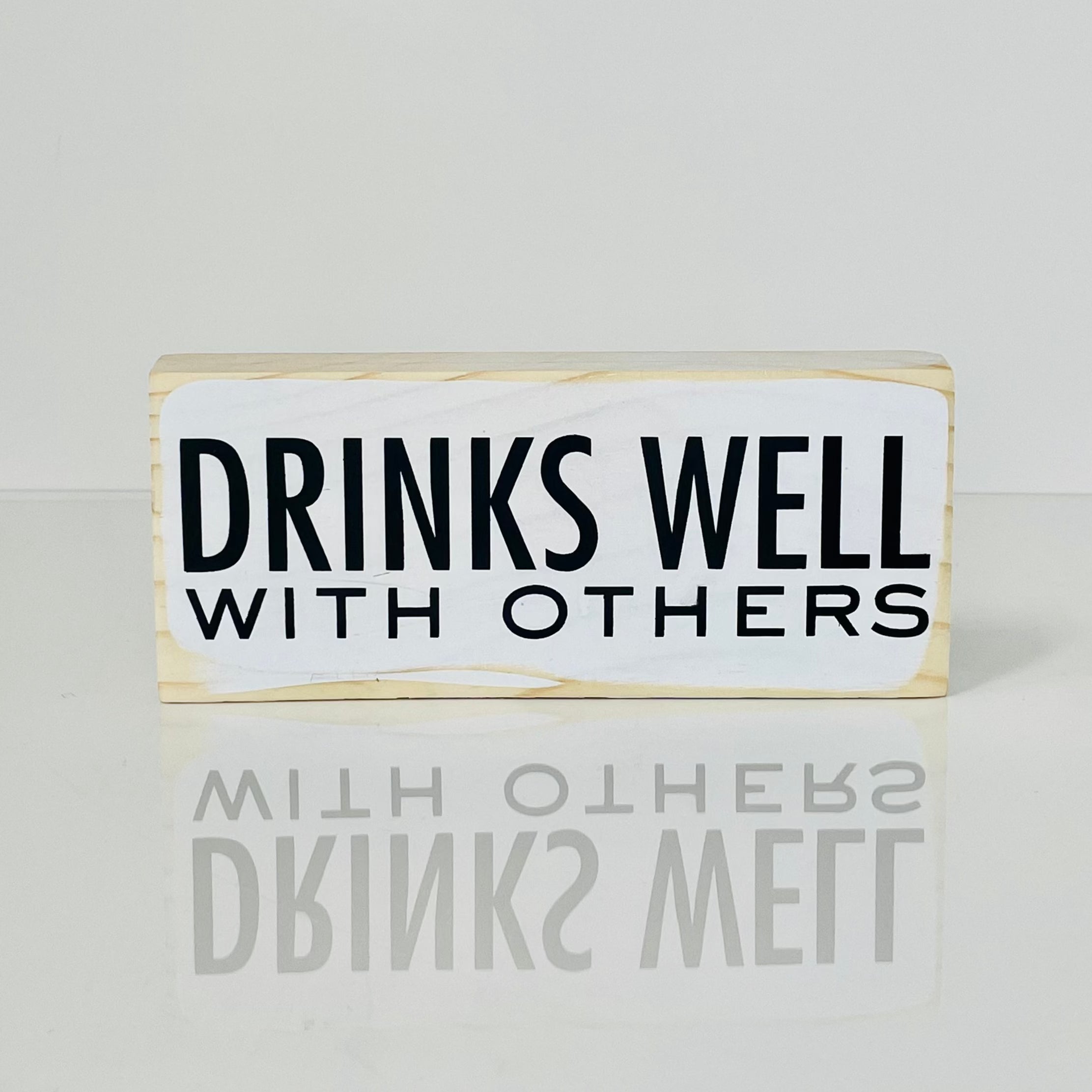 a small, white, wood sign sits on a white surface. It reads, in black, "Drinks well with others"