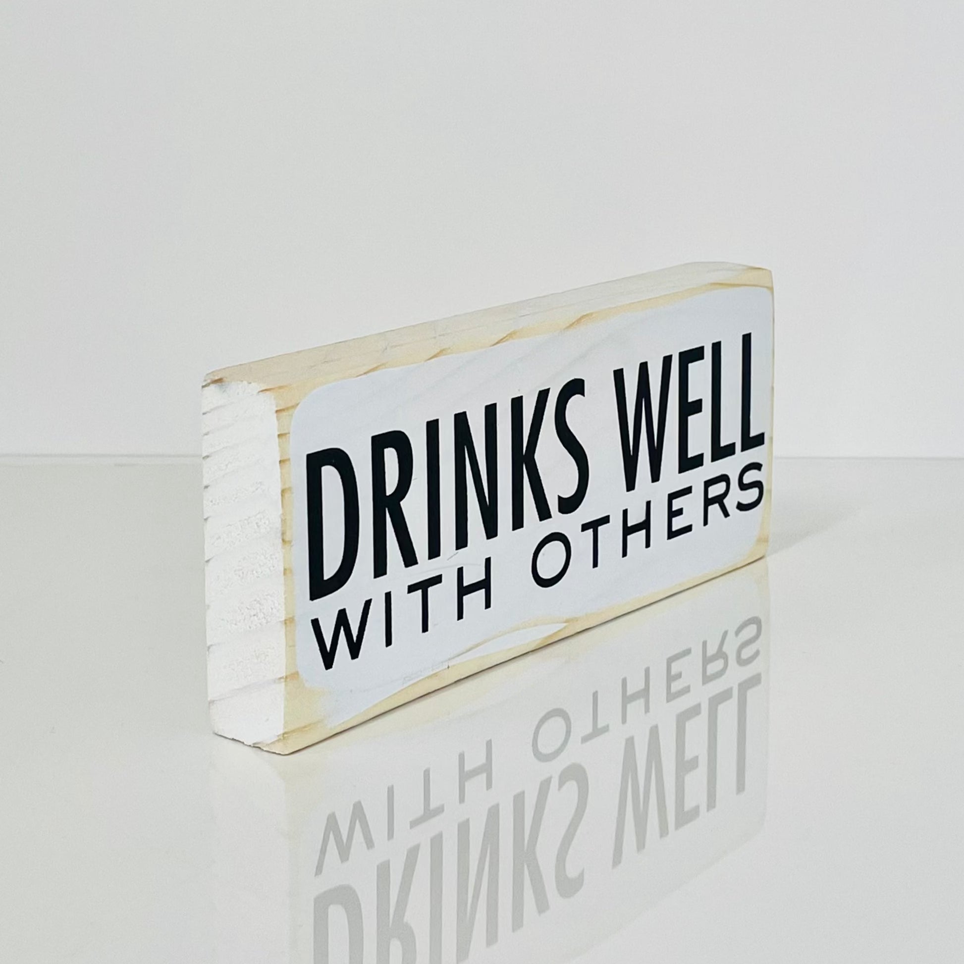 side view of a small, white, wood sign sits on a white surface. It reads, in black, "Drinks well with others"