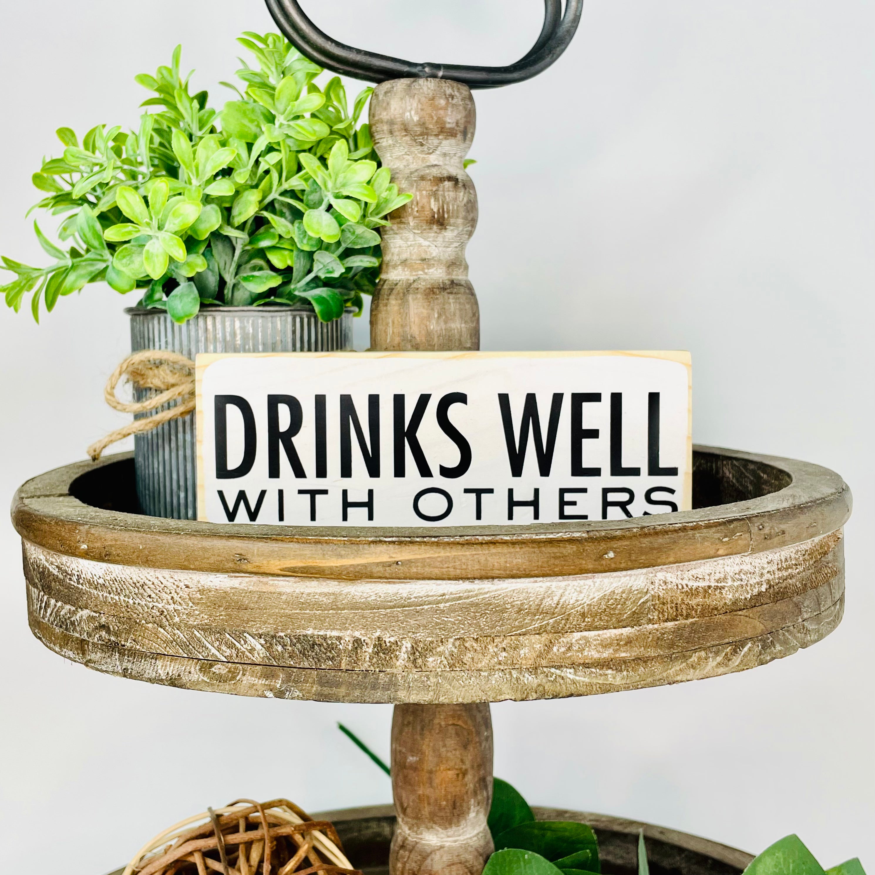 a small, white, wood, sign sits on a tiered tray. It reads, in black, "drinks well with others".