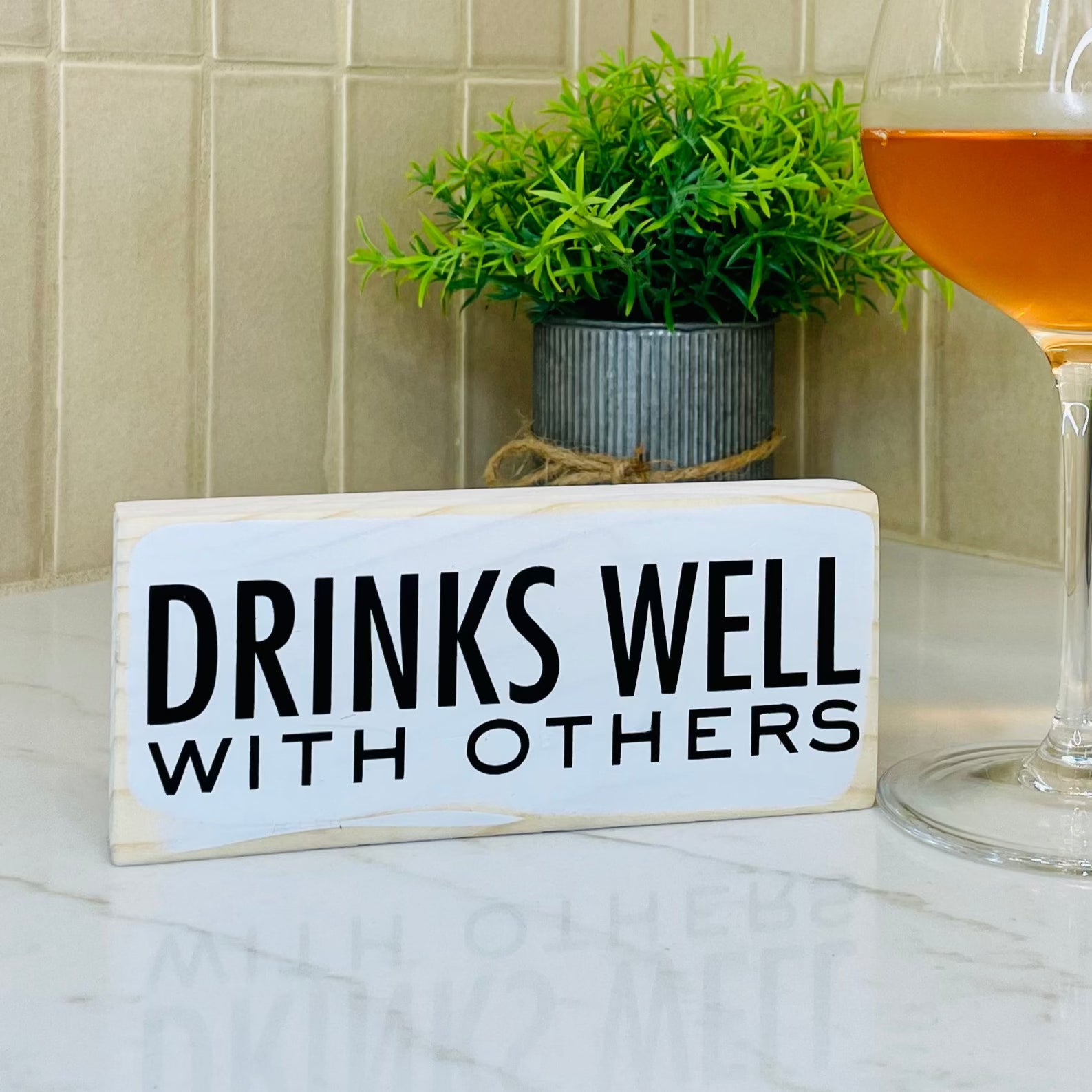 a small, white, wood sign sits on a kitchen counter next to a glass of wine. The sign reads, in black, "Drinks well with others".