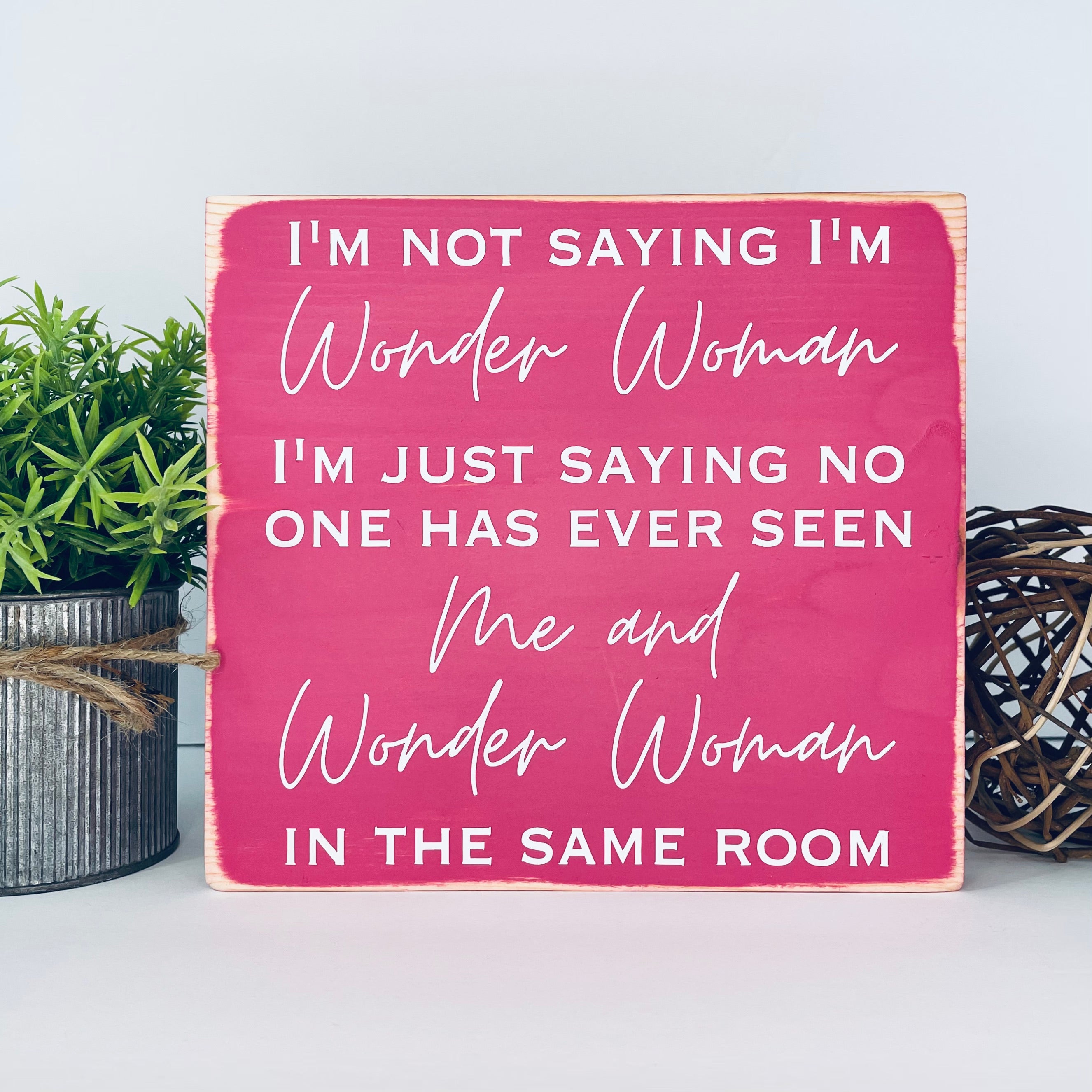 medium sized square wood sign painted pink with white lettering that reads, " I'm not saying I'm Wonder Woman, I'm just saying no one has ever seen me and Wonder Woman in the same room". All text is in all caps except for "Wonder Woman" and "Me and Wonder Woman". Text is centered, takes up the entire area and is on 7 lines. Glossy finish.