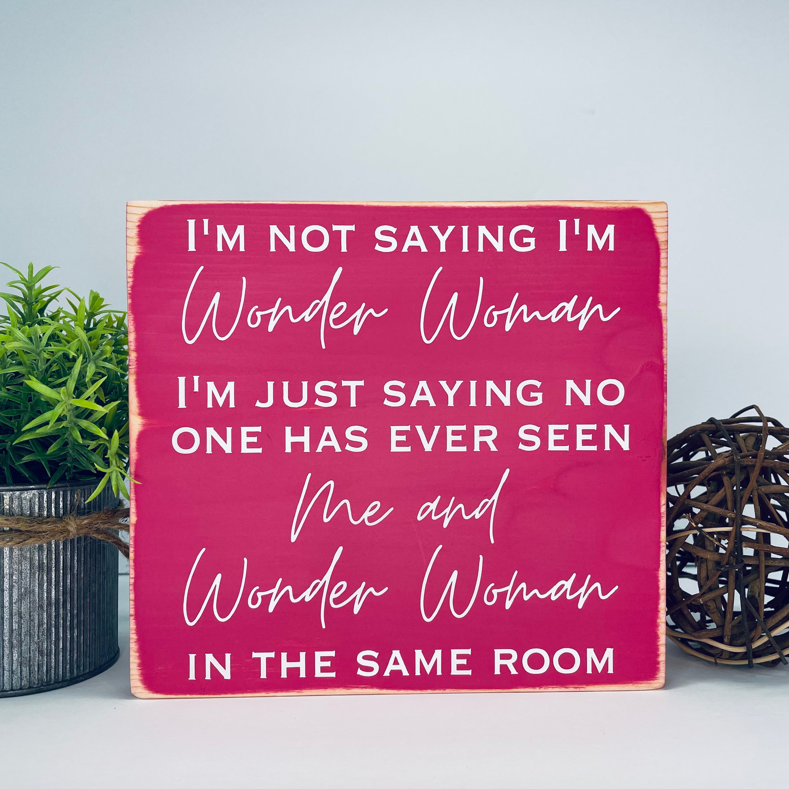 medium sized square wood sign painted pink with white lettering that reads, " I'm not saying I'm Wonder Woman, I'm just saying no one has ever seen me and Wonder Woman in the same room". All text is in all caps except for "Wonder Woman" and "Me and Wonder Woman". Text is centered, takes up the entire area and is on 7 lines. Glossy finish.