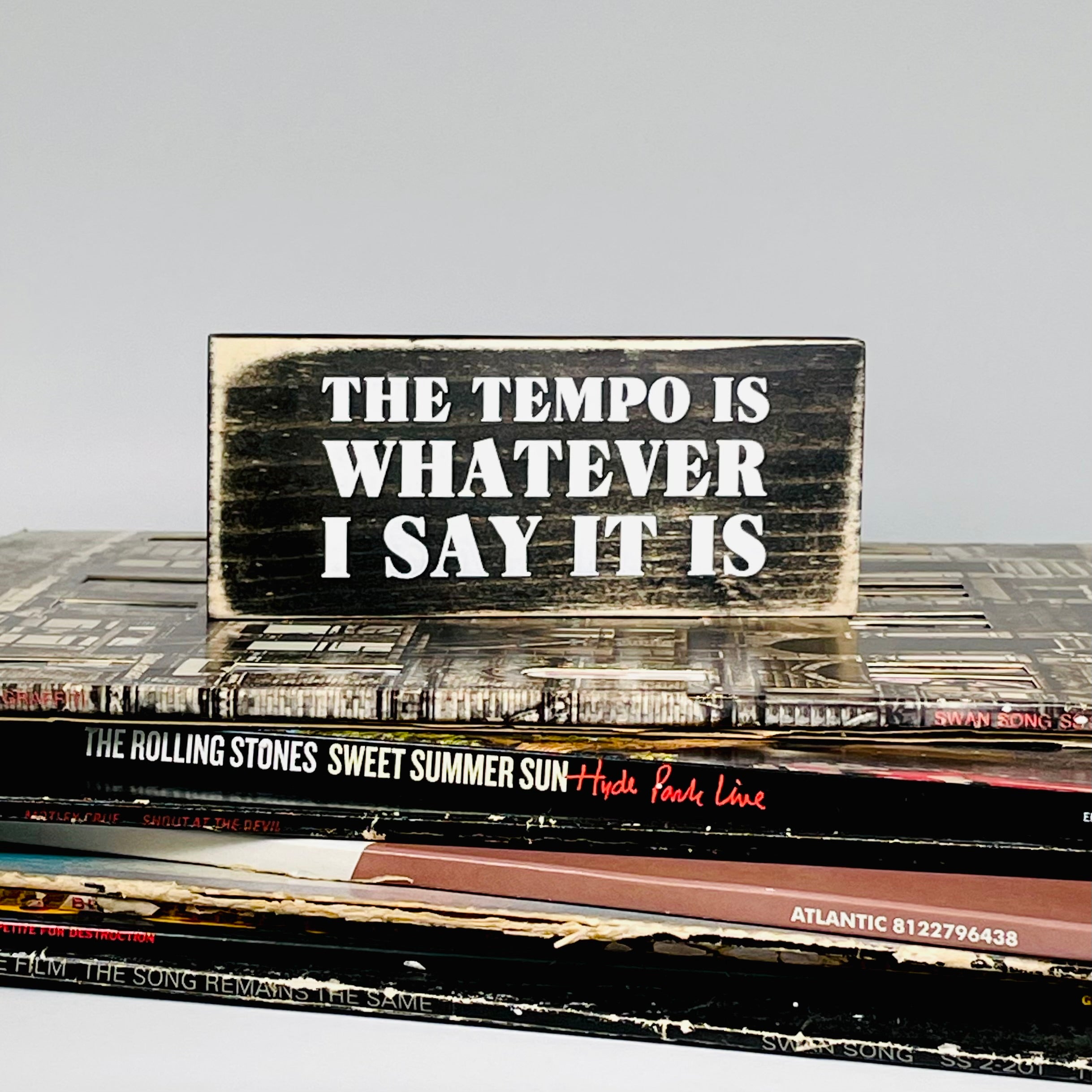A small, black, wood sign with white lettering is sitting on a stack of vinyl records. In white the sign reads, "THE TEMPO IS WHATEVER I SAY IT IS"