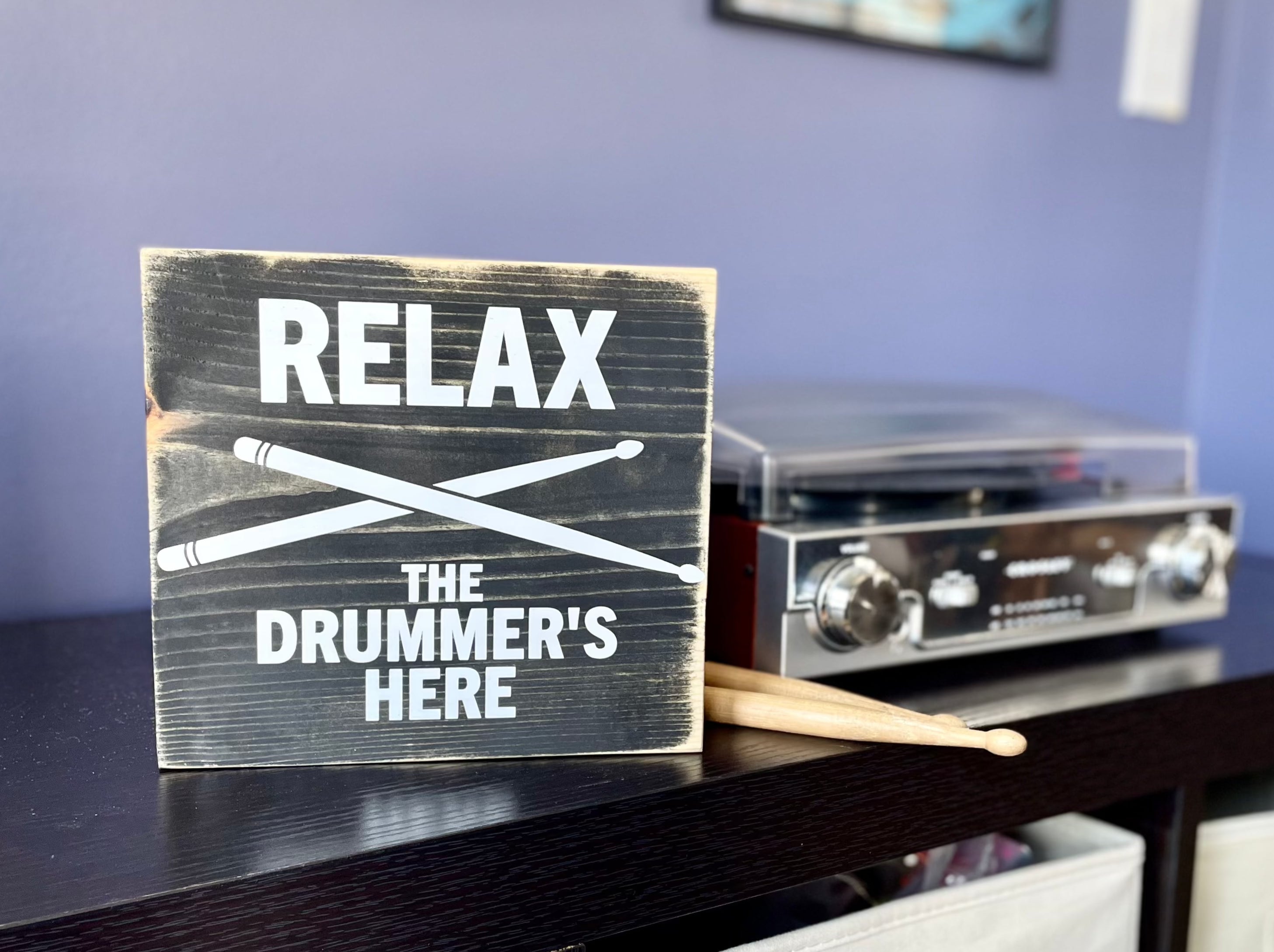 A black, wood sign with white lettering is sitting on a black shelf next to a record player and a set of wood drumsticks. The sign reads: "Relax the Drummer's Here." A white pair of drumsticks is painted in an x under the word relax.