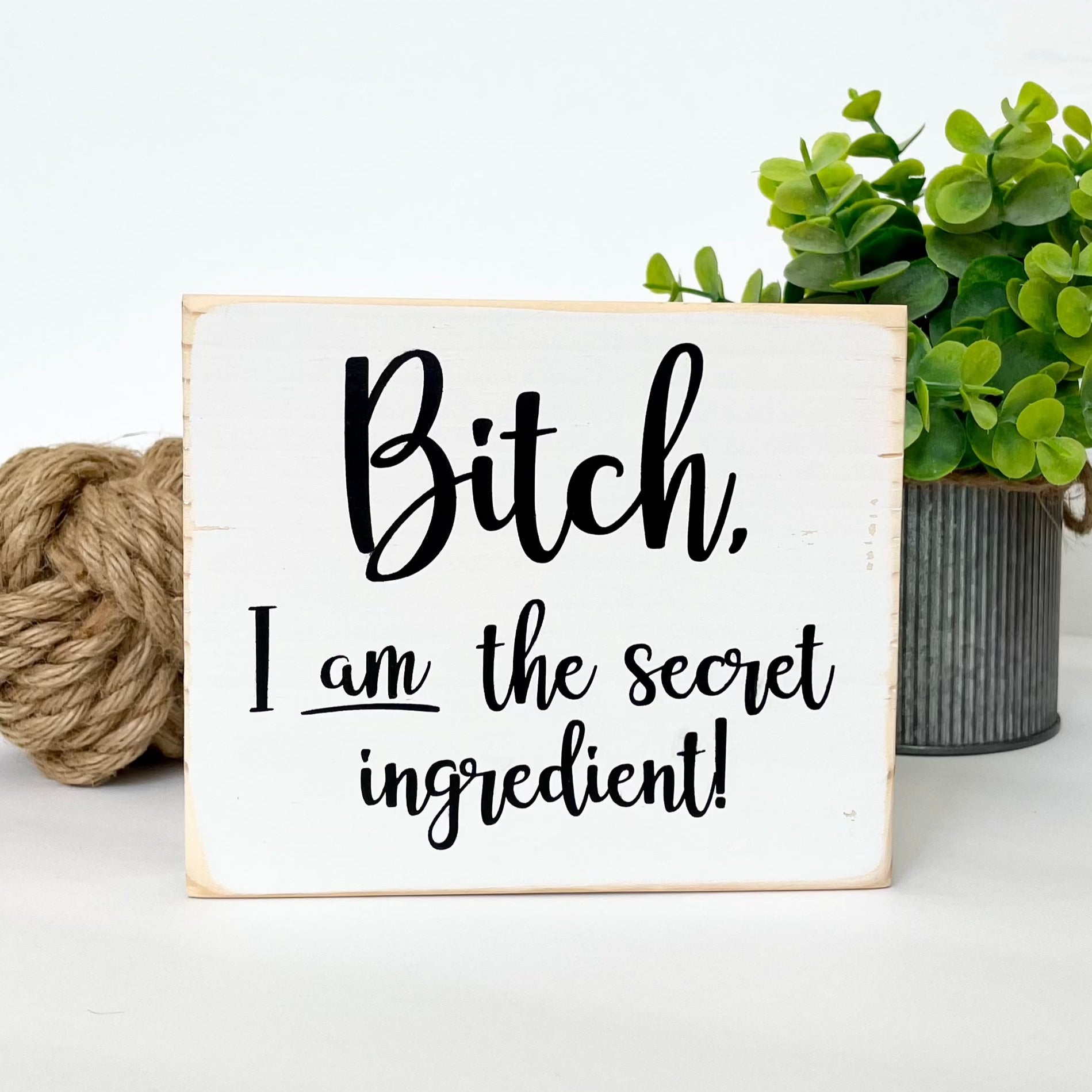 A small, white, wood sign sits on a white surface. The sign says, in black, "Bitch, I am the secret ingredient".