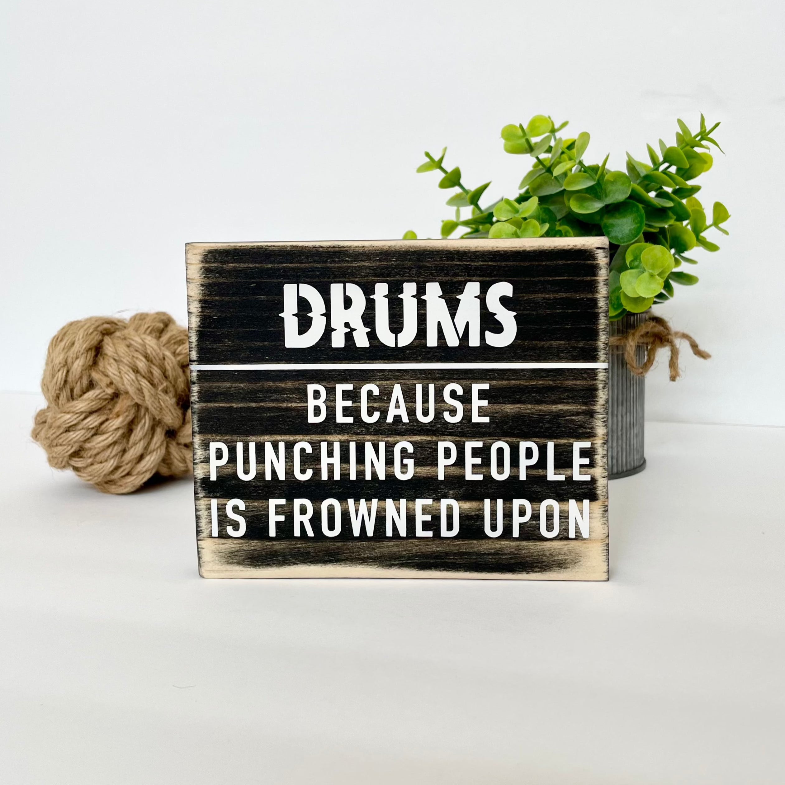 A small, black, wood sign sits on a white table next to a small plant. In white it reads, "Drums. Because punching people is frowned upon."