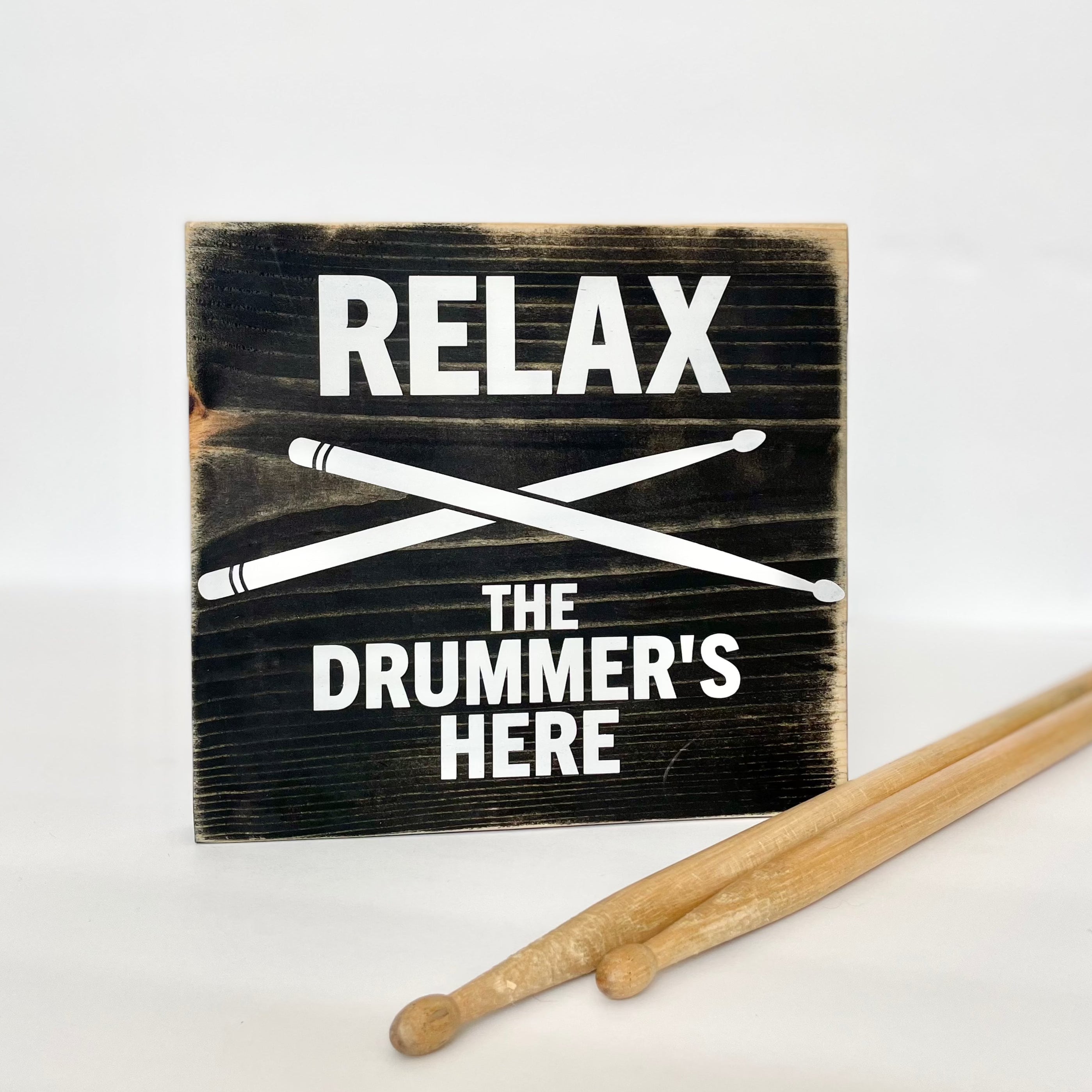 A black, wood sign with white lettering sits on a white shelf next to a pair of wood drumsticks. The sign reads: "Relax the Drummer's Here." A white pair of drumsticks is painted in an x under the word relax.