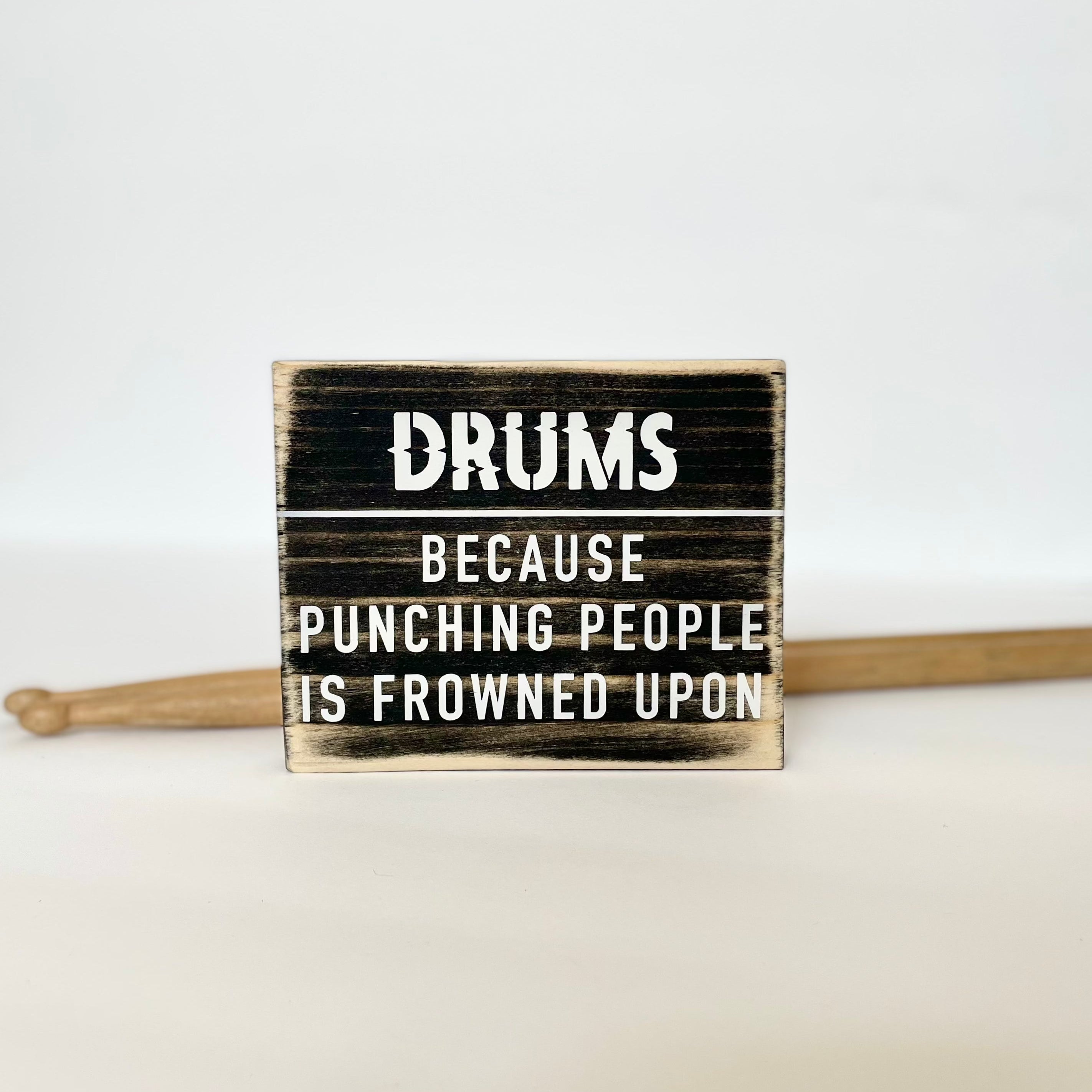 A small, black, wood sign sits on a white table next to a pair of drumsticks. In white it reads, "Drums. Because punching people is frowned upon."