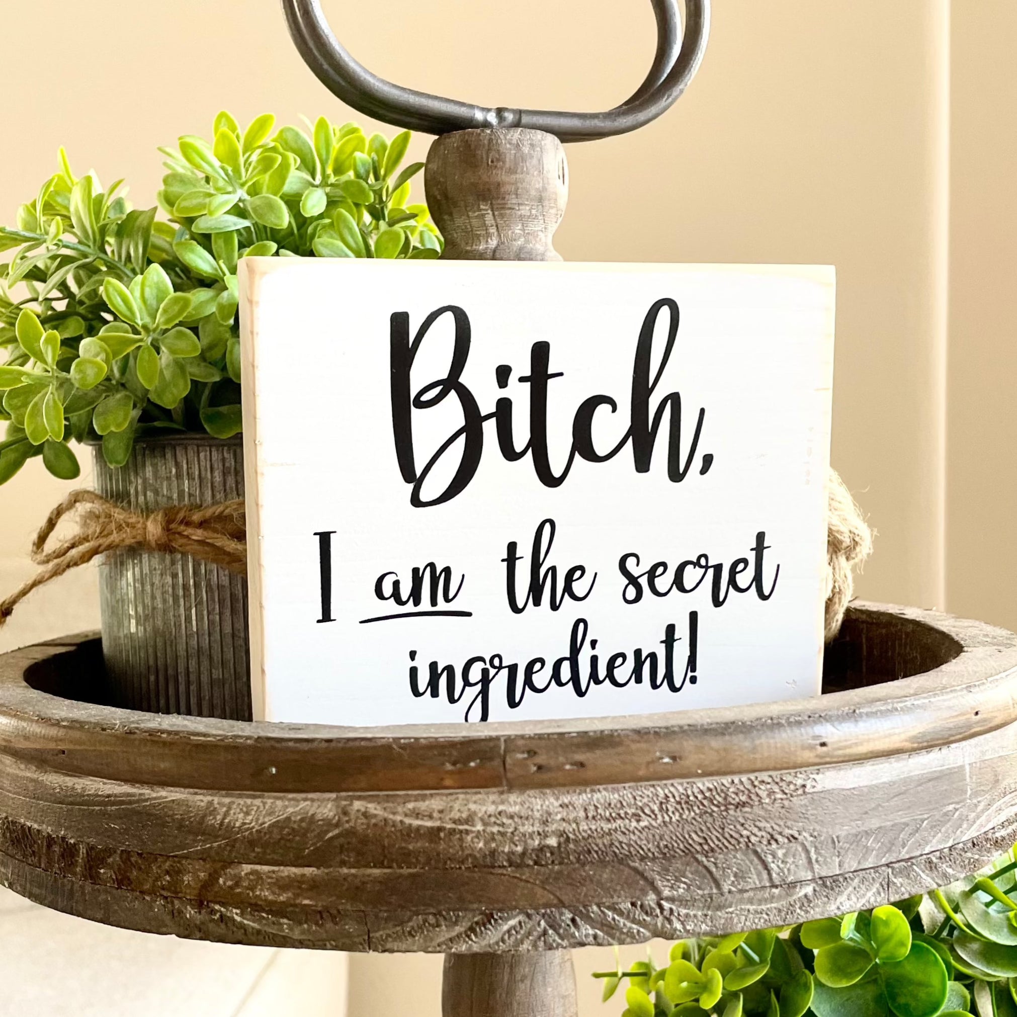 A small, white, wood sign sits on a tiered tray. The sign says, in black, "Bitch, I am the secret ingredient".