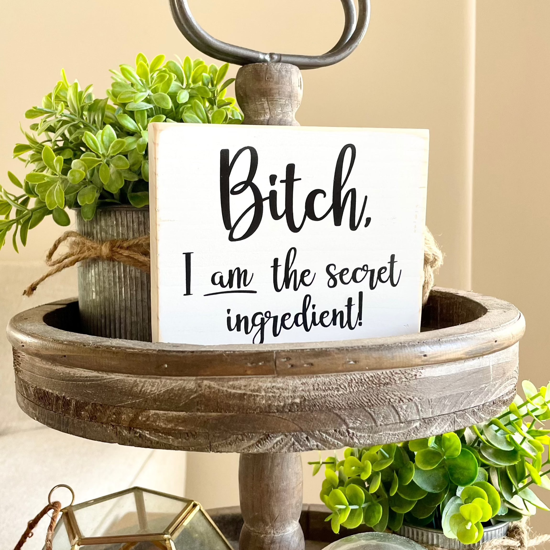 A small, white, wood sign sits on a tiered tray. The sign says, in black, "Bitch, I am the secret ingredient".