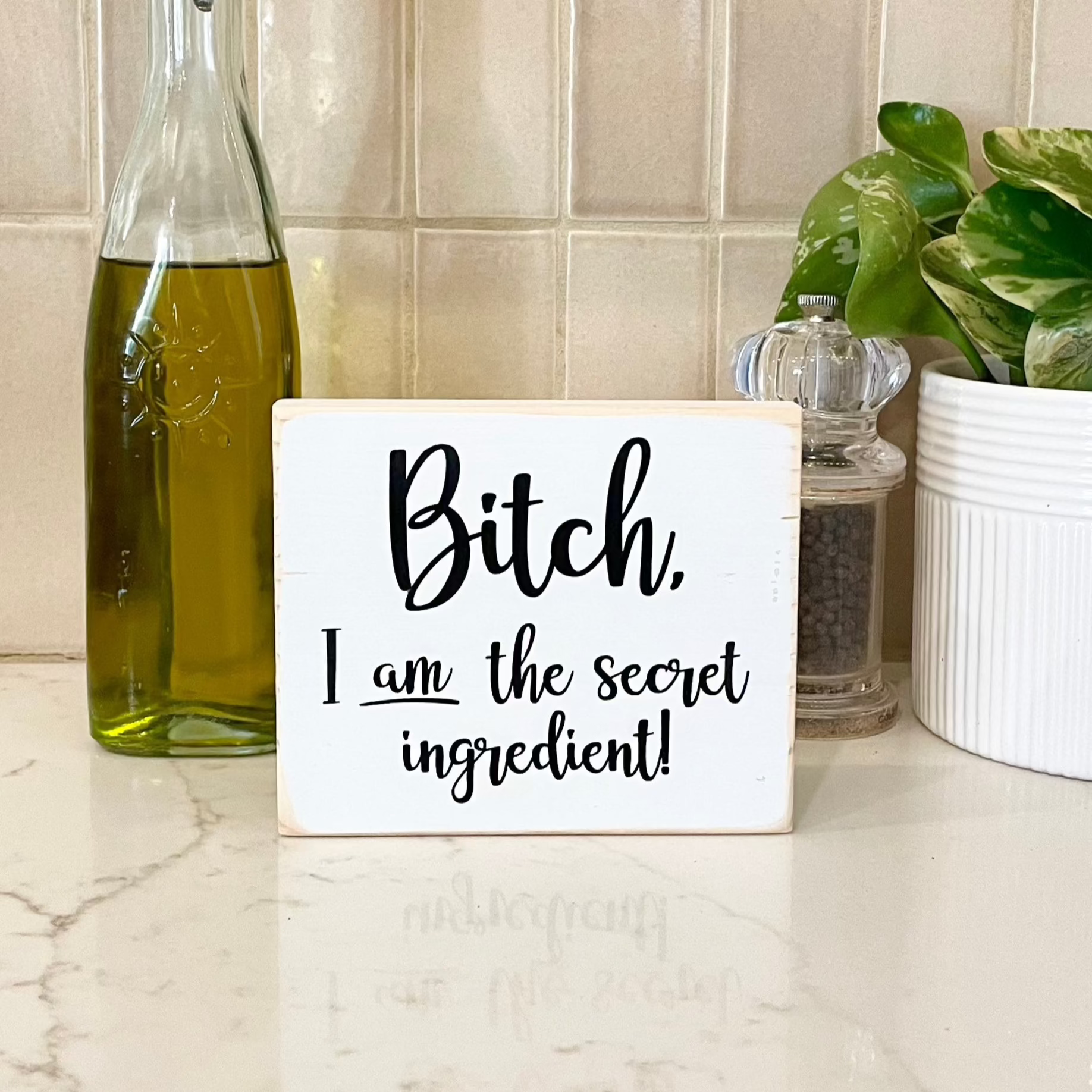 A small, white, wood sign sits on a kitchen counter with olive oil and a pepper grinder. The sign says, in black, "Bitch, I am the secret ingredient".