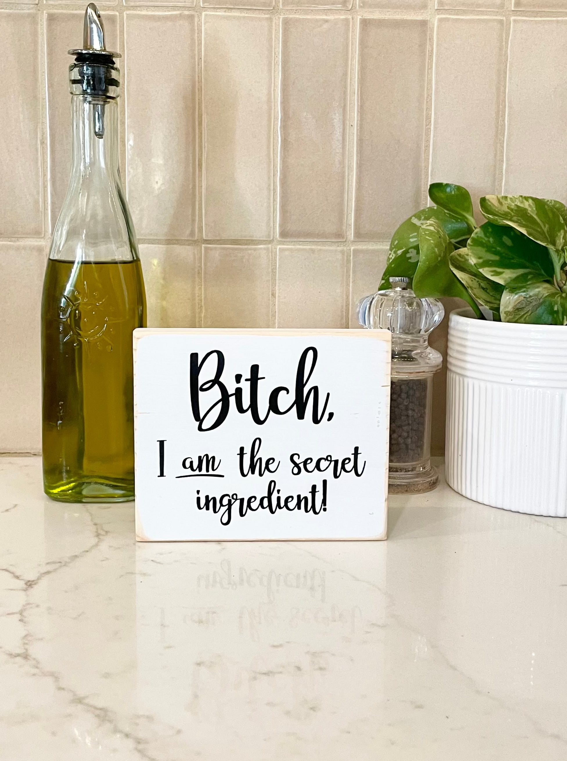 A small, white, wood sign sits on a kitchen counter with olive oil and a pepper grinder. The sign says, in black, "Bitch, I am the secret ingredient".