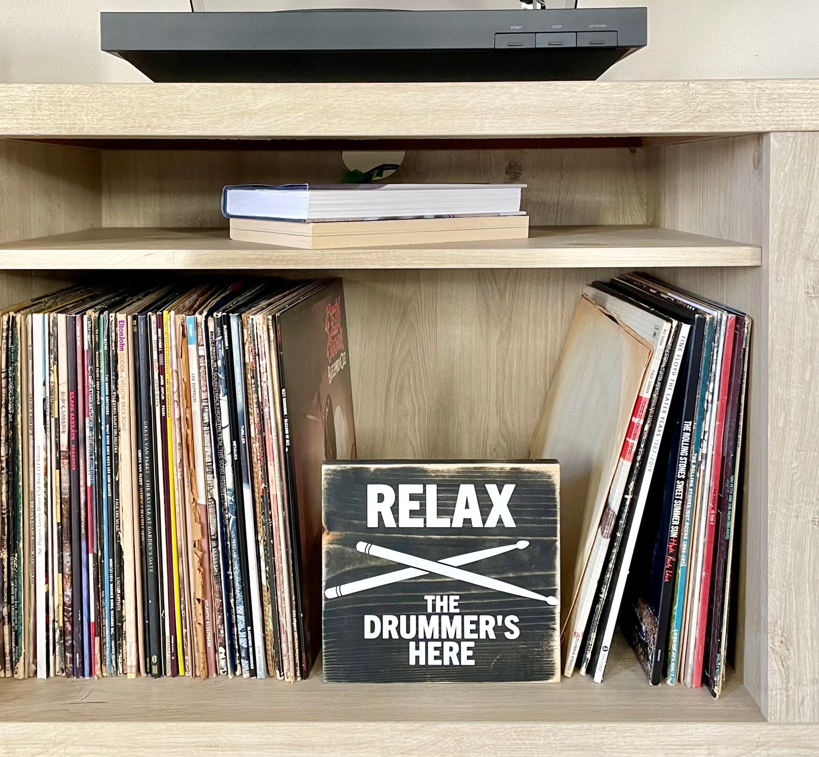 A black, wood sign with white lettering sits on a shelf next to a stack of vinyl records. The sign reads: "Relax the Drummer's Here." A white pair of drumsticks is painted in an x under the word relax.