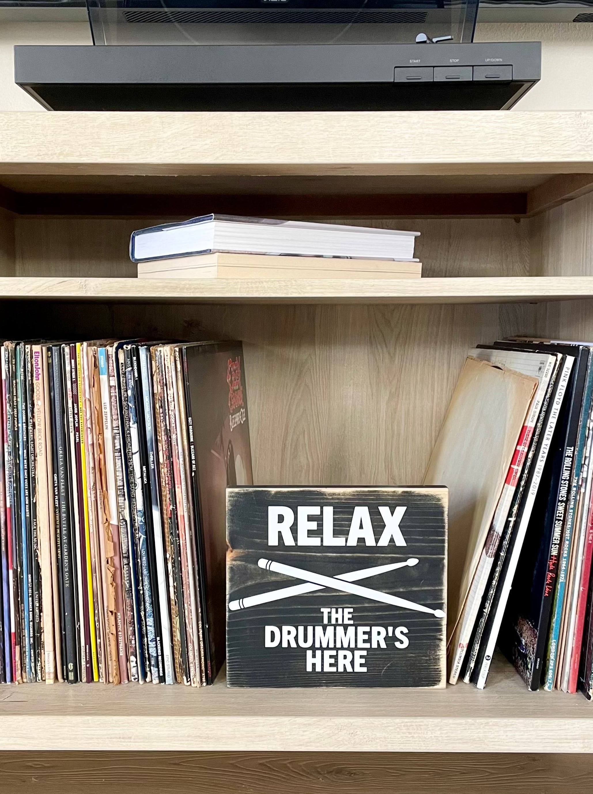 A black, wood sign with white lettering sits on a shelf next to a stack of vinyl records. . The sign reads: "Relax the Drummer's Here." A white pair of drumsticks is painted in an x under the word relax.