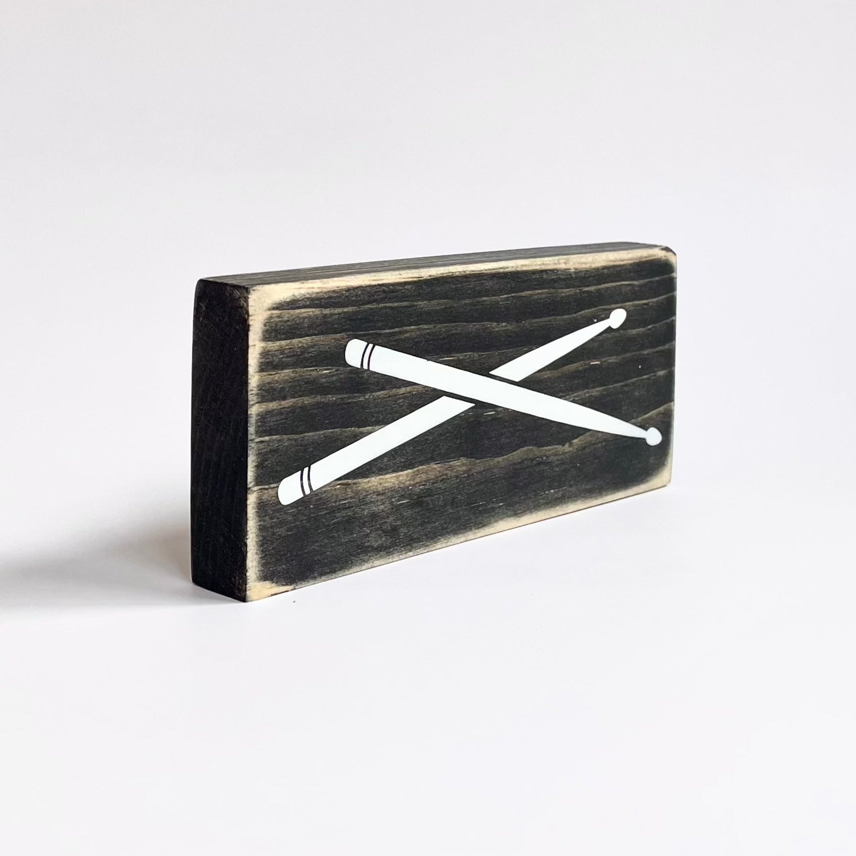 Side view of a small, black, wood sign with drumsticks painted on it, sits horizontally on a white surface..