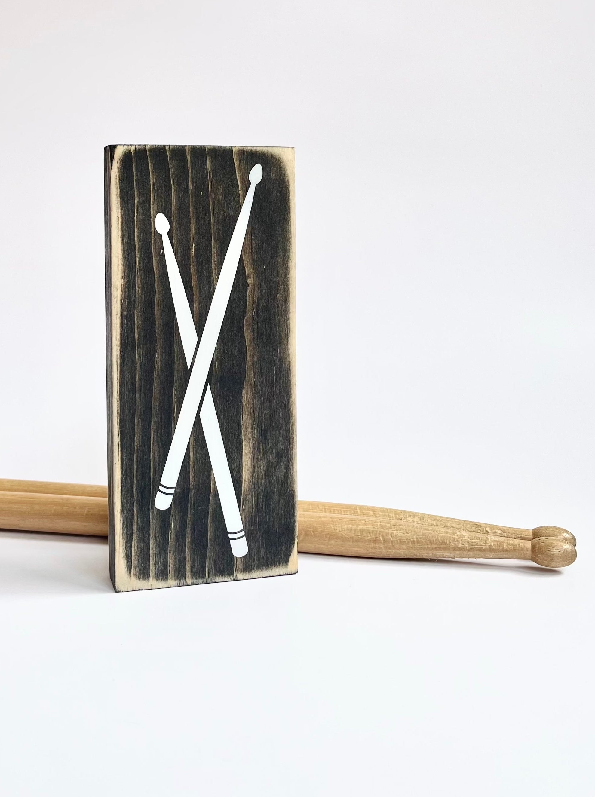 A small, black, wood sign with drumsticks painted on it, sits vertically on a white surface.