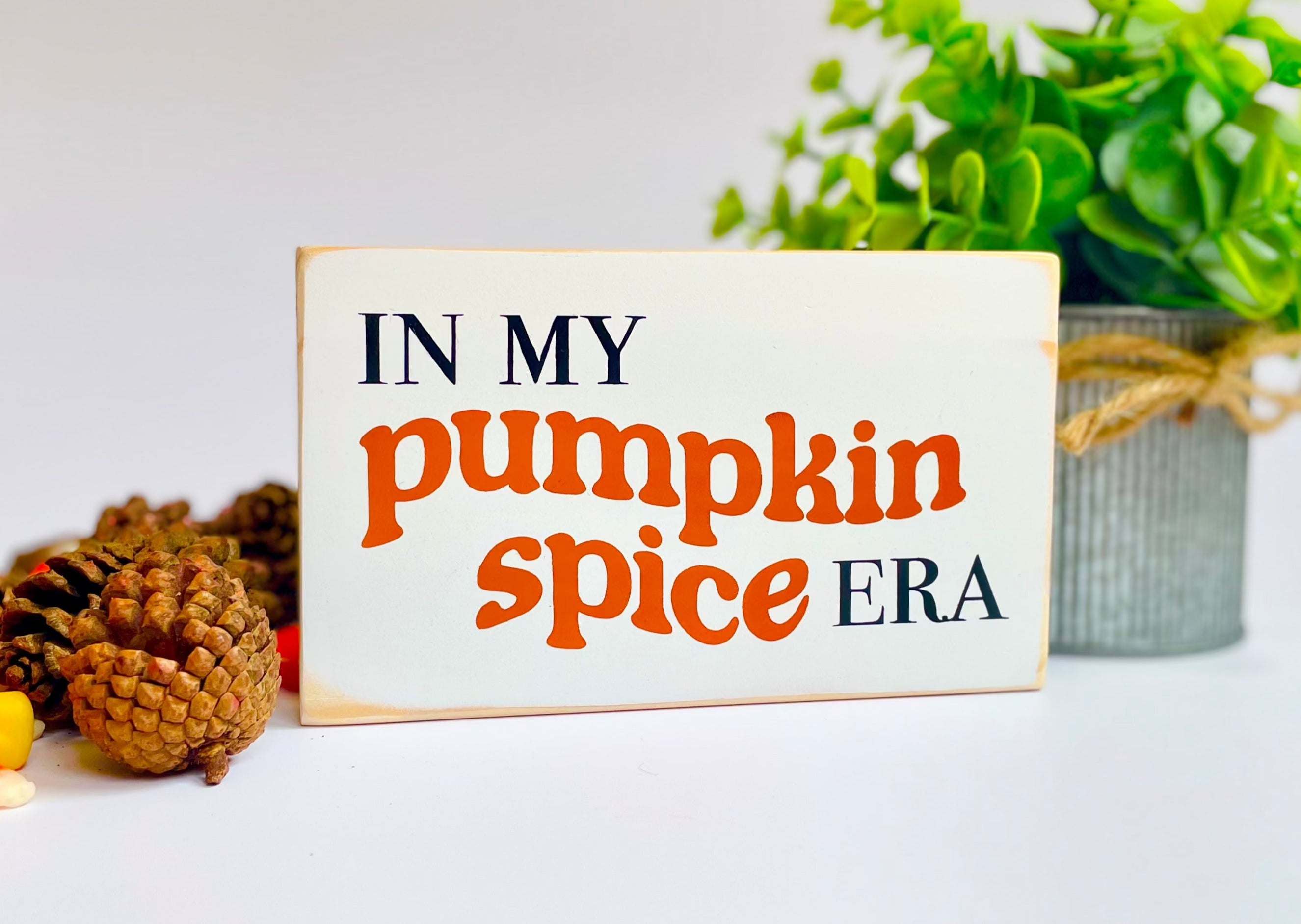 a small, white, wood sign that reads" in my pumpkin spice era" sits on a white surface with a pinecone and a plant next to it.