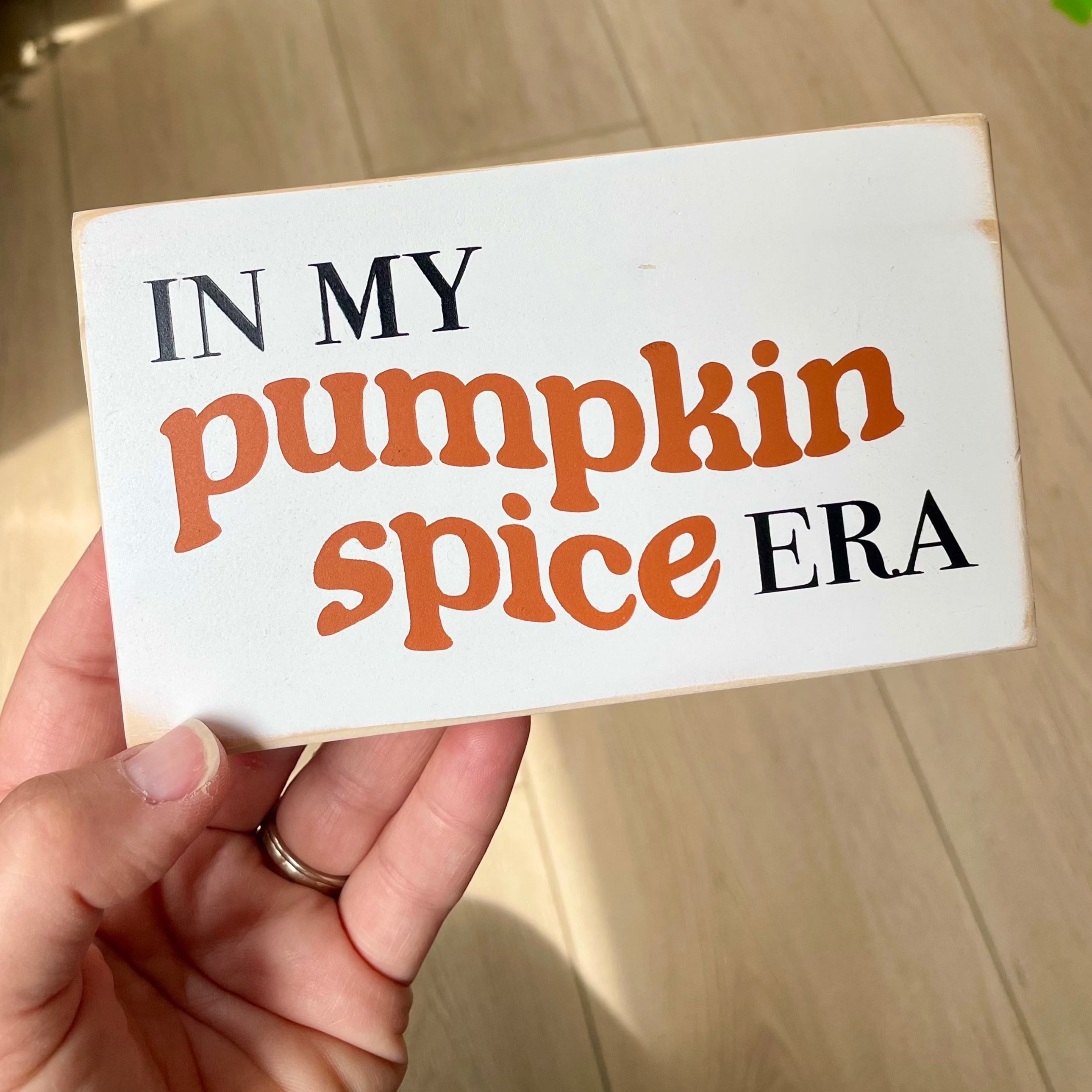 a small, white, wood sign that reads" in my pumpkin spice era" is being held in a person's hand with a blurred background