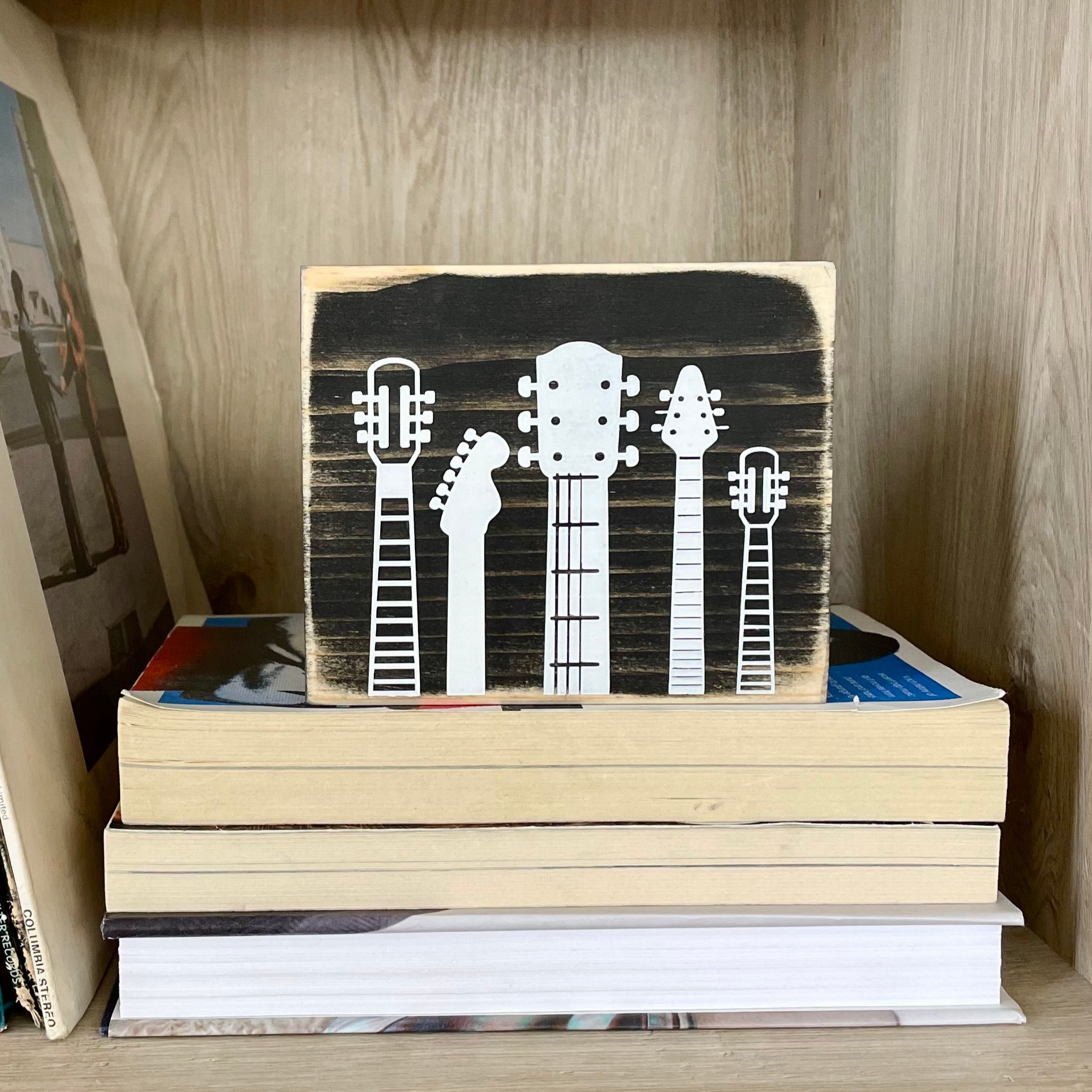 A small, rustic, black, wood sign with white guitar heads on it, sits on a shelf on a stack of books.