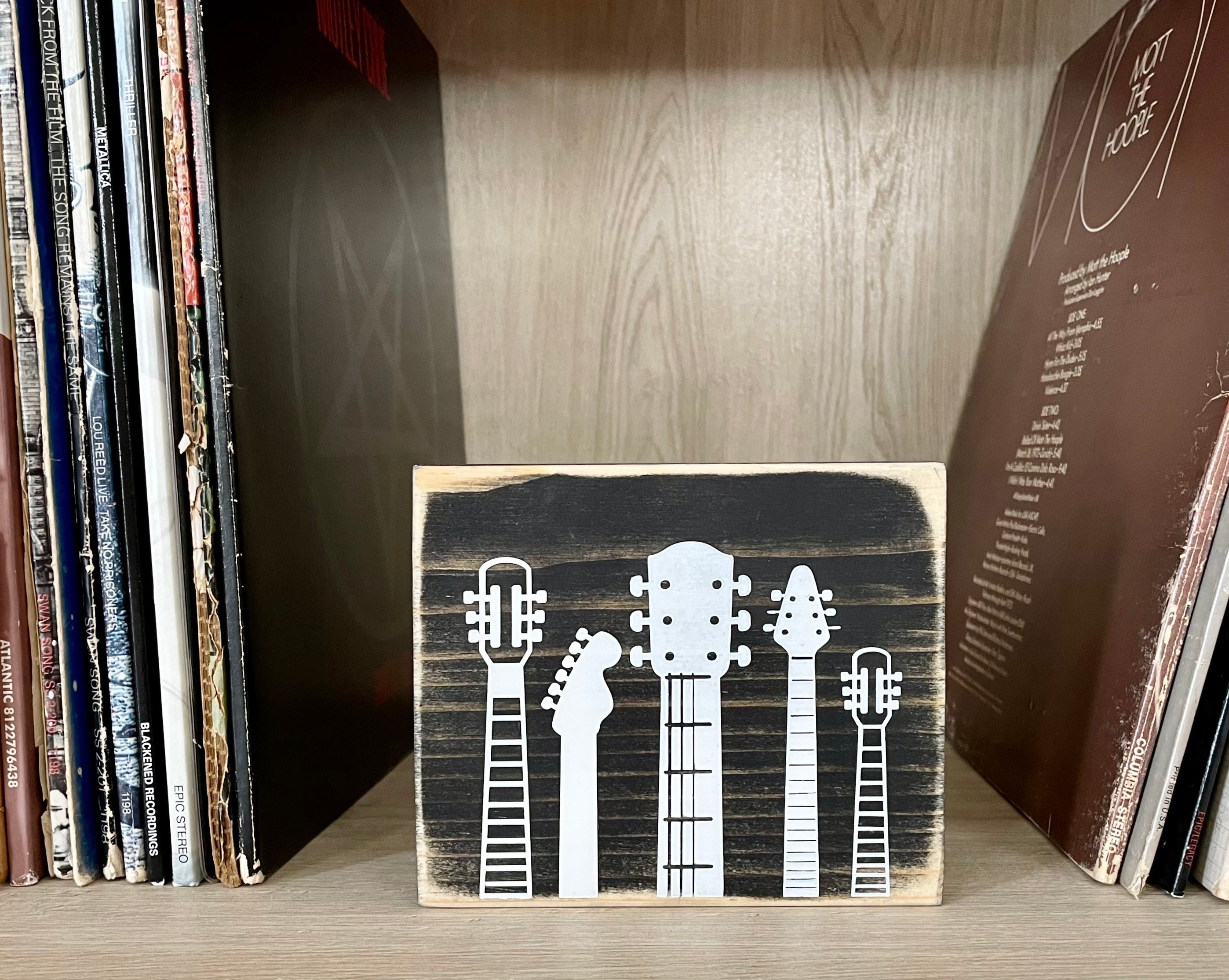 A small, rustic, black, wood sign with white guitar heads on it, sits on a shelf next to vinyl records.