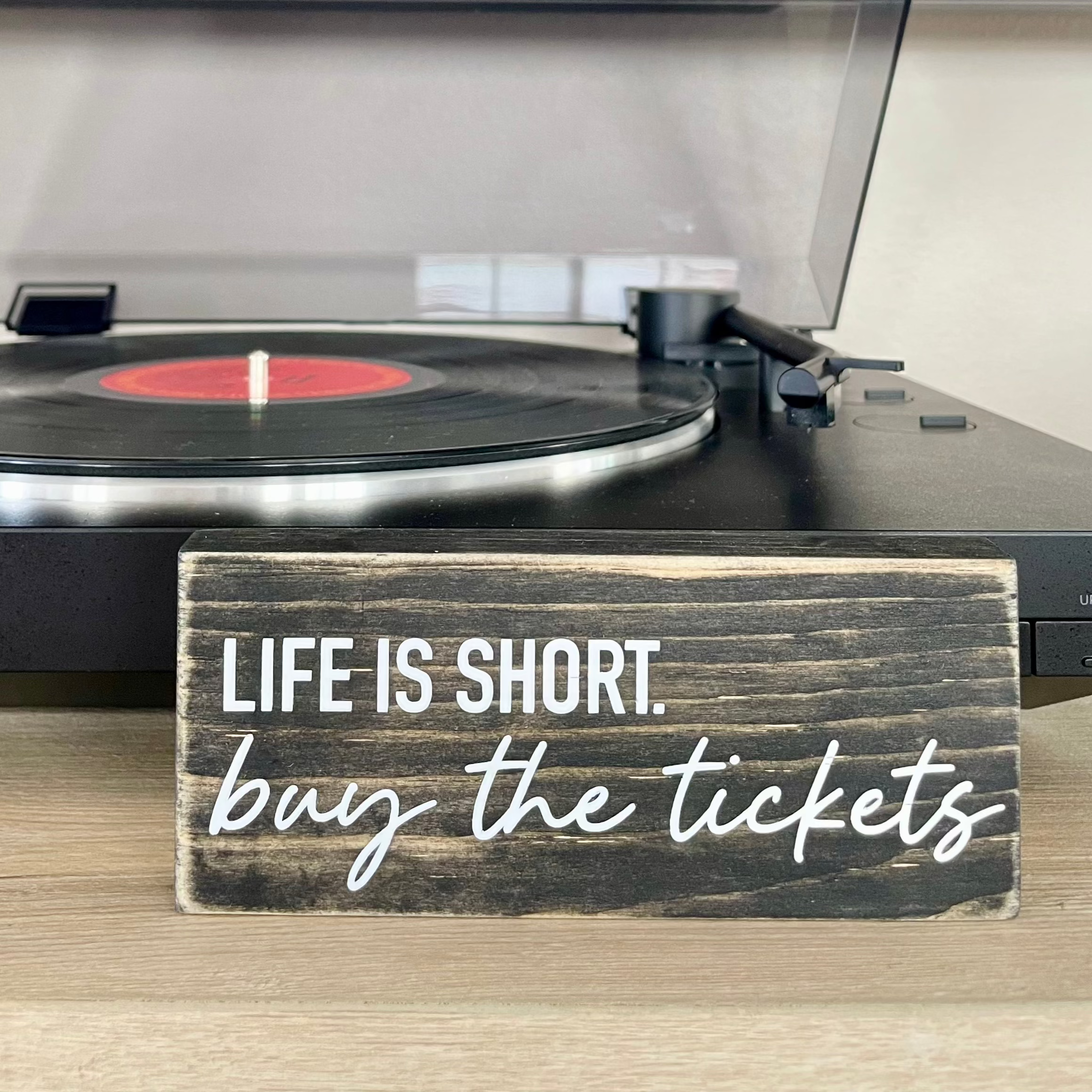 a small, black, wood sign sits on a table in front of a record player. The sign reads, in white, "Life is short. Buy the tickets"