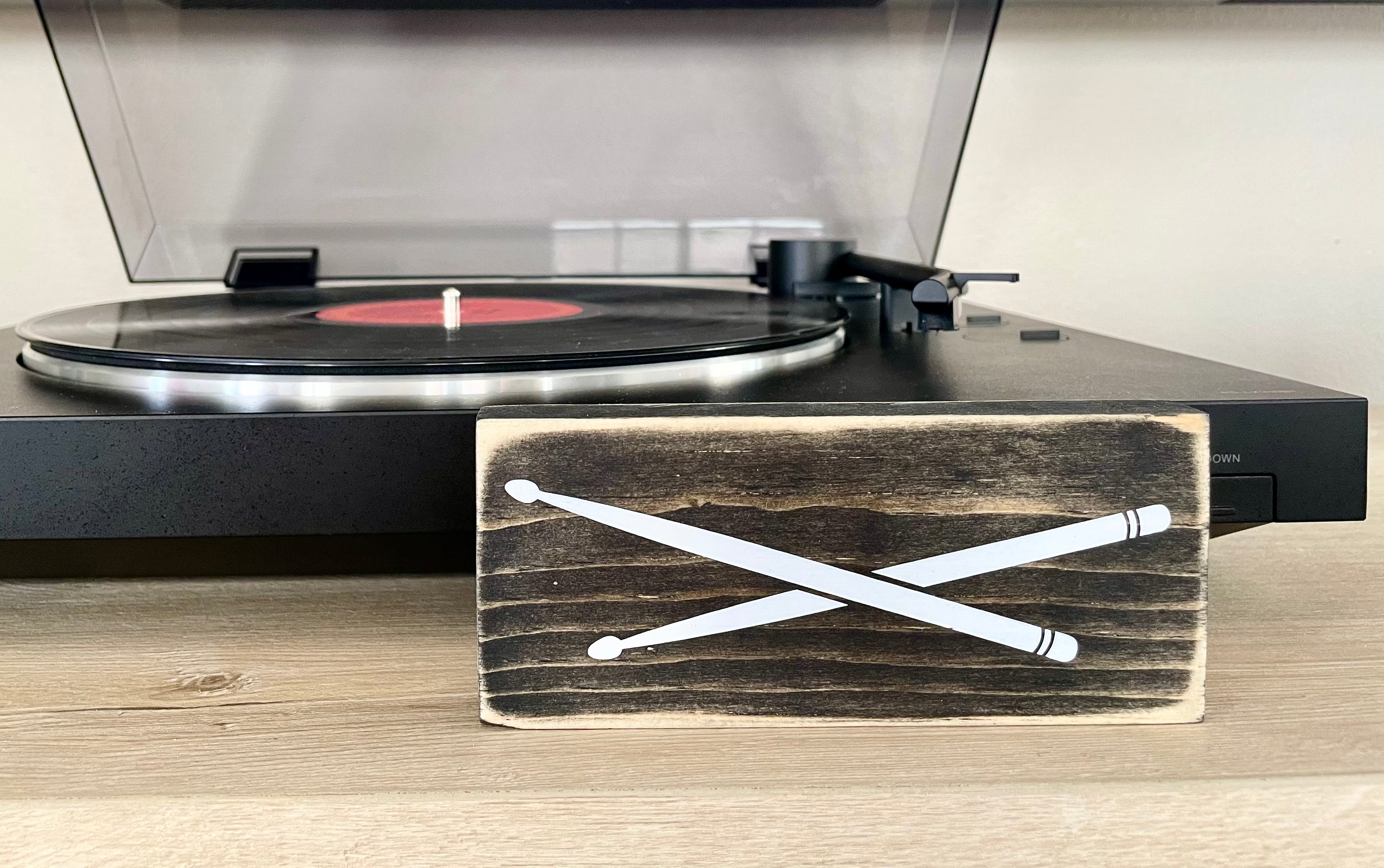 A small, black, wood sign with drumsticks painted on it, sits horizontally on a table in front of a record player.