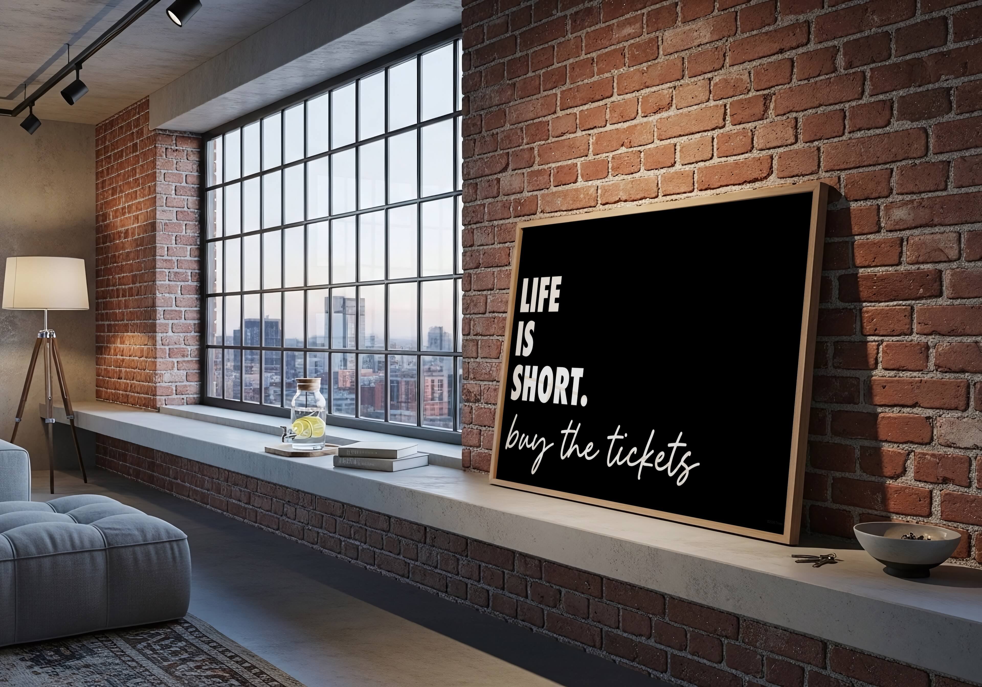 A framed black sign with white text reading 'LIFE IS SHORT. buy the tickets' leaning against a red brick wall in a modern loft-style room with a large window and exposed brick.