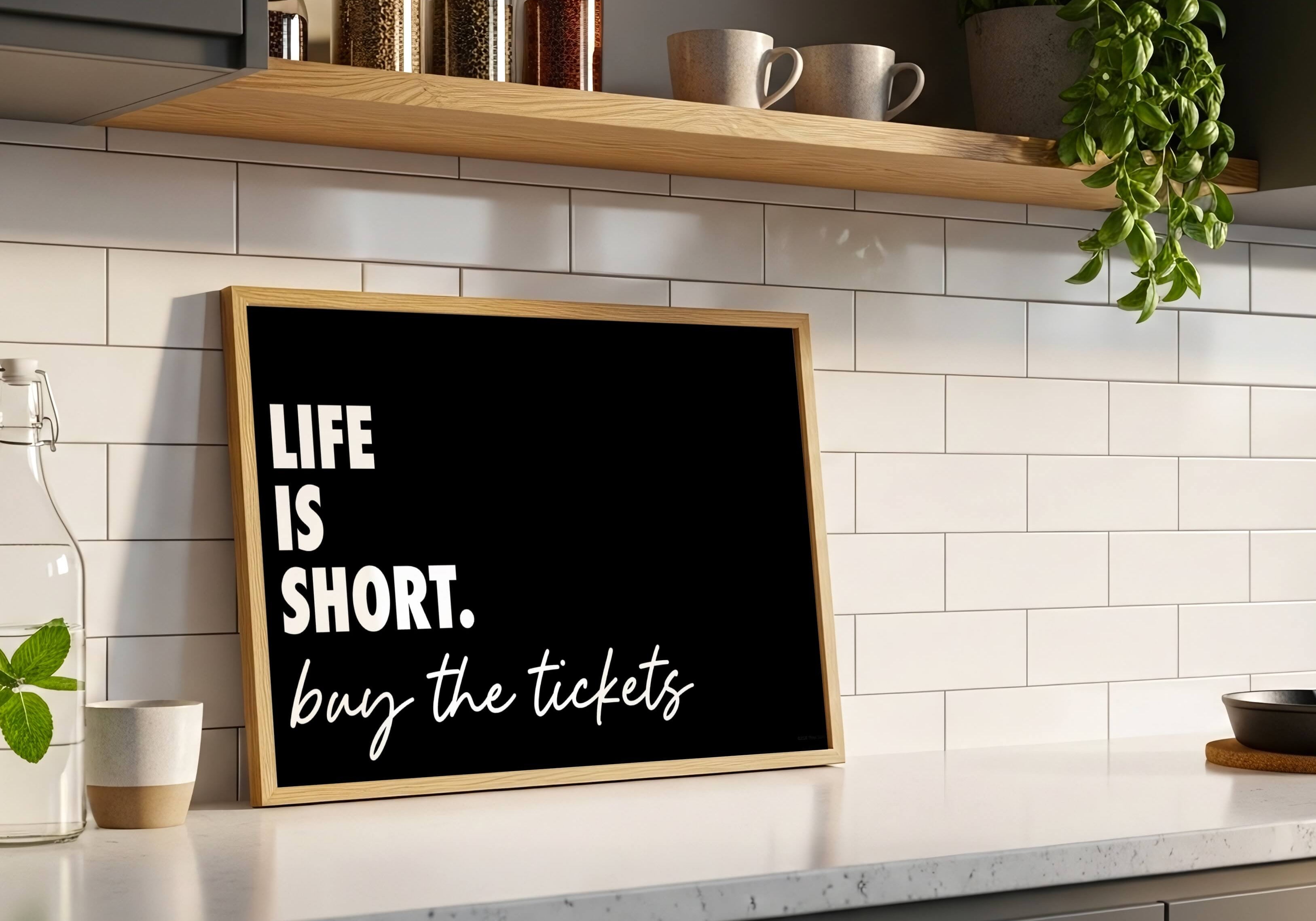 A framed black print with the quote 'LIFE IS SHORT. buy the tickets' in white lettering, leaning on a kitchen counter against a white subway tile backsplash.