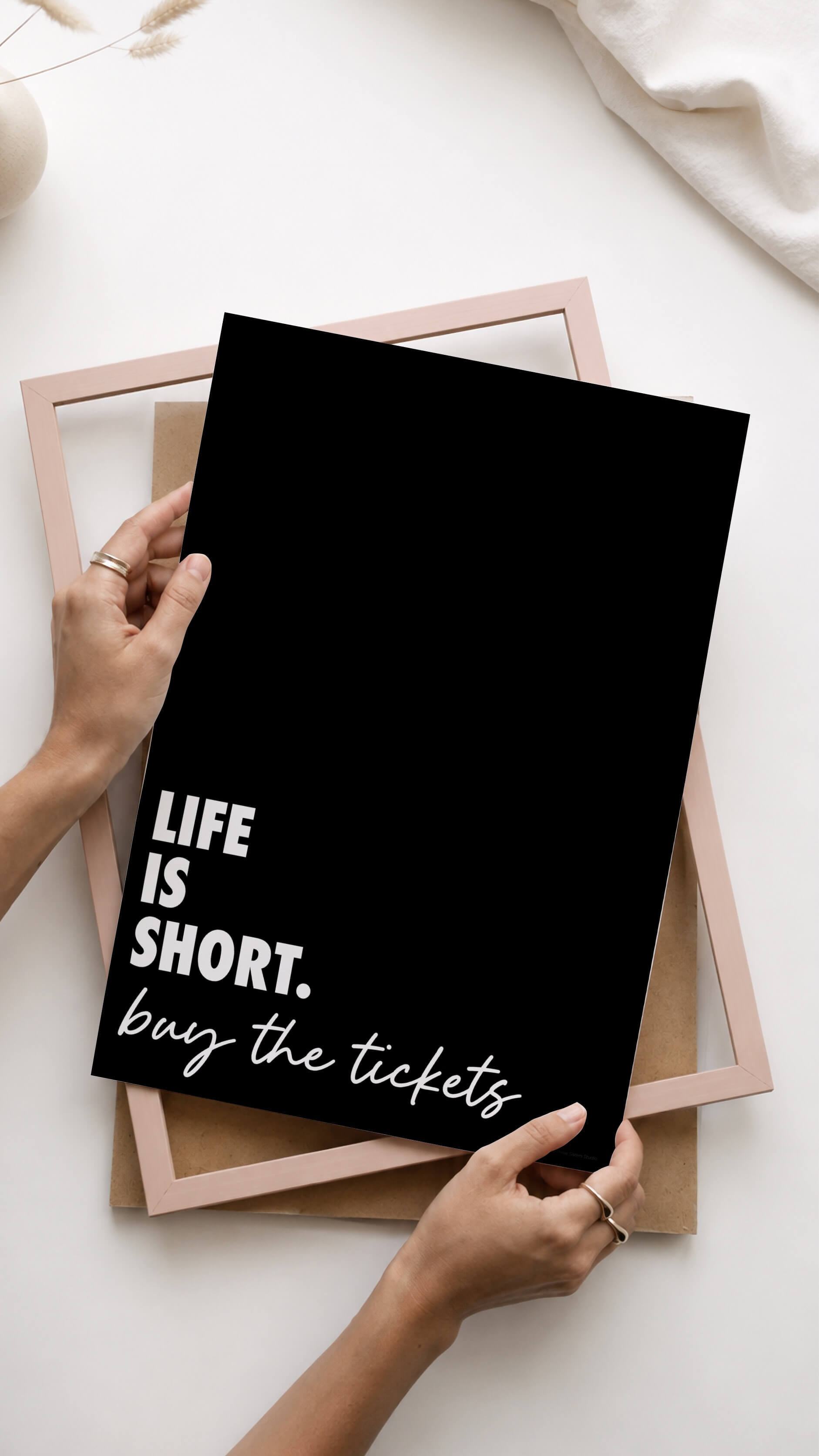 A person holds a black art print with white text reading 'LIFE IS SHORT. buy the tickets' over a stack of pastel pink and brown picture frames on a white surface.