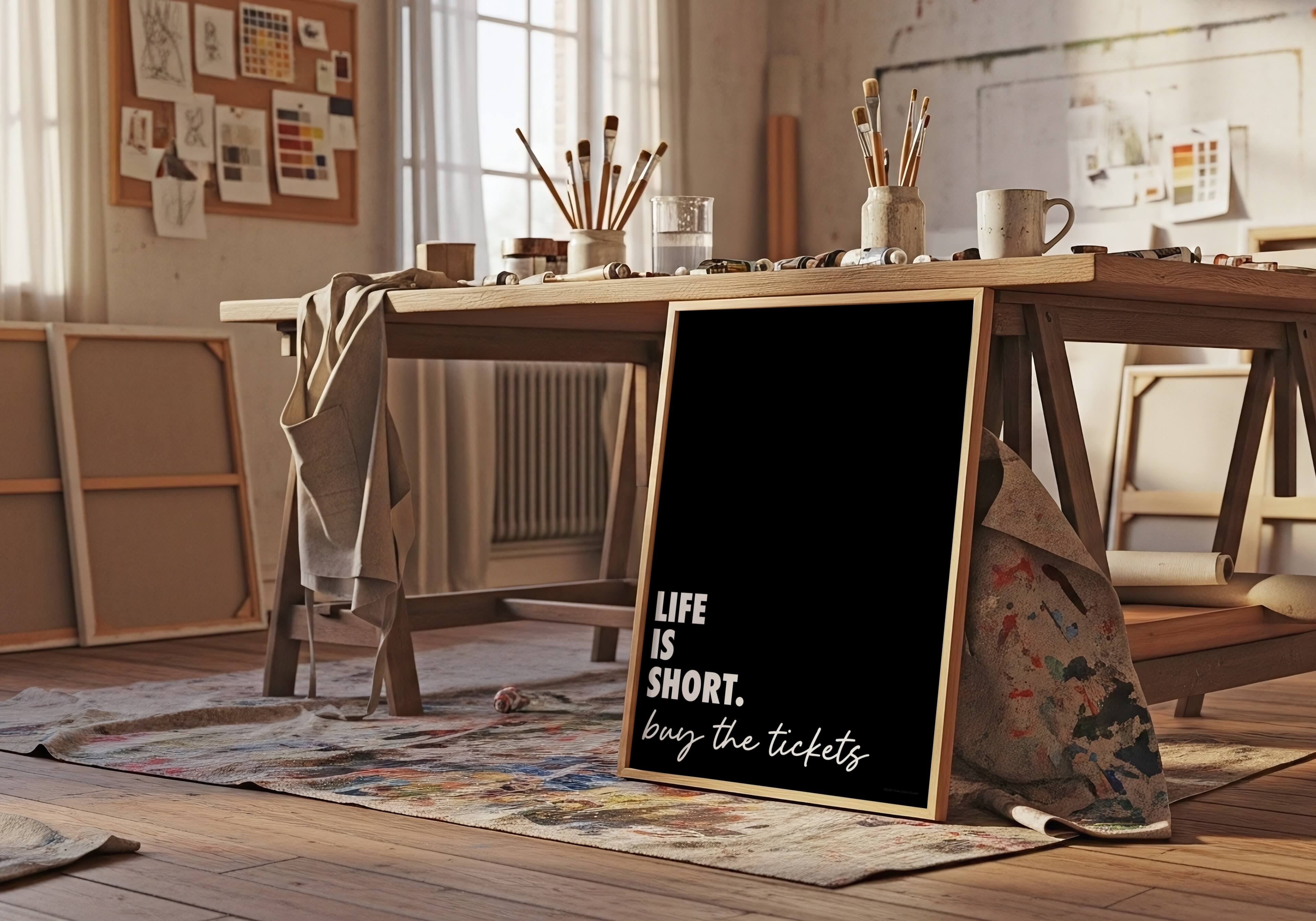 A framed black sign with white text reading 'LIFE IS SHORT. buy the tickets' stands on a floor in a warm, sunlit artist studio.