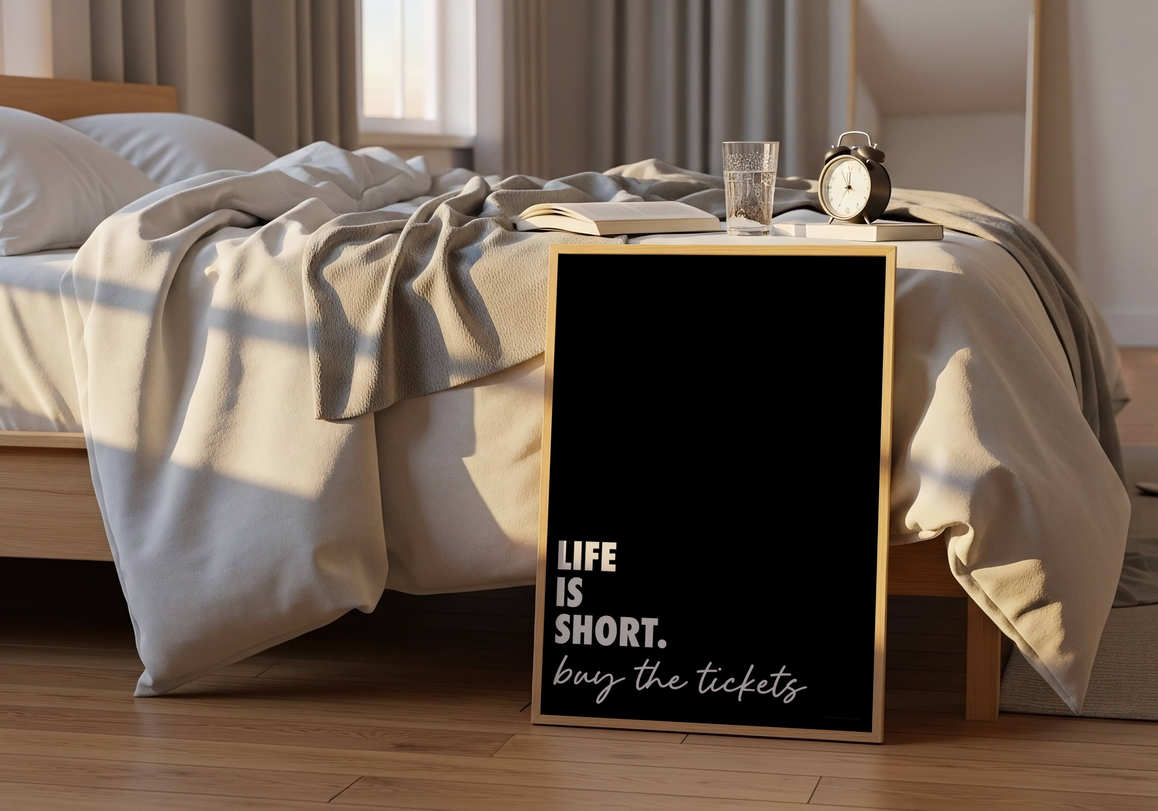 A framed black sign with white text reading 'LIFE IS SHORT. buy the tickets' stands on a wooden floor beside an unmade bed in a softly lit bedroom.