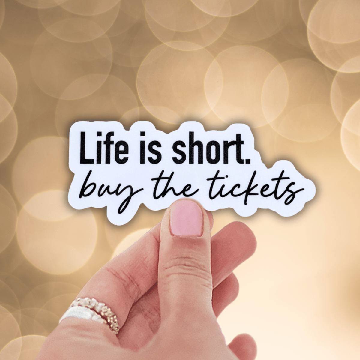 Hand holding a sticker with 'Life is short, buy the tickets' text against a blurred background.