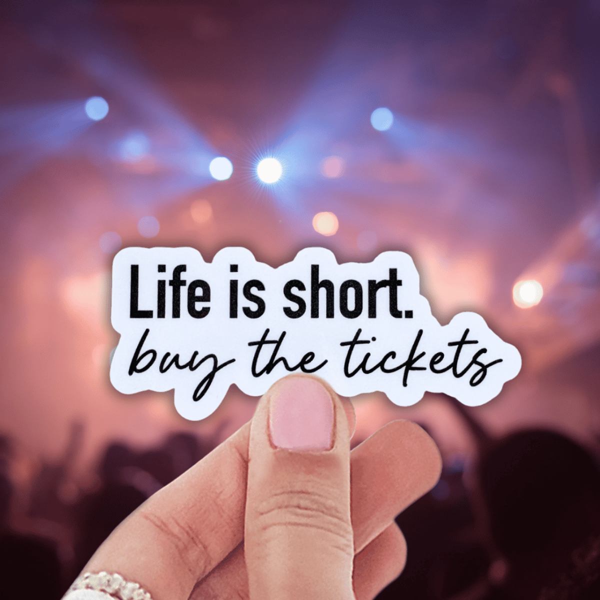 Hand holding a 'Life is short, buy the tickets' sticker with a concert background