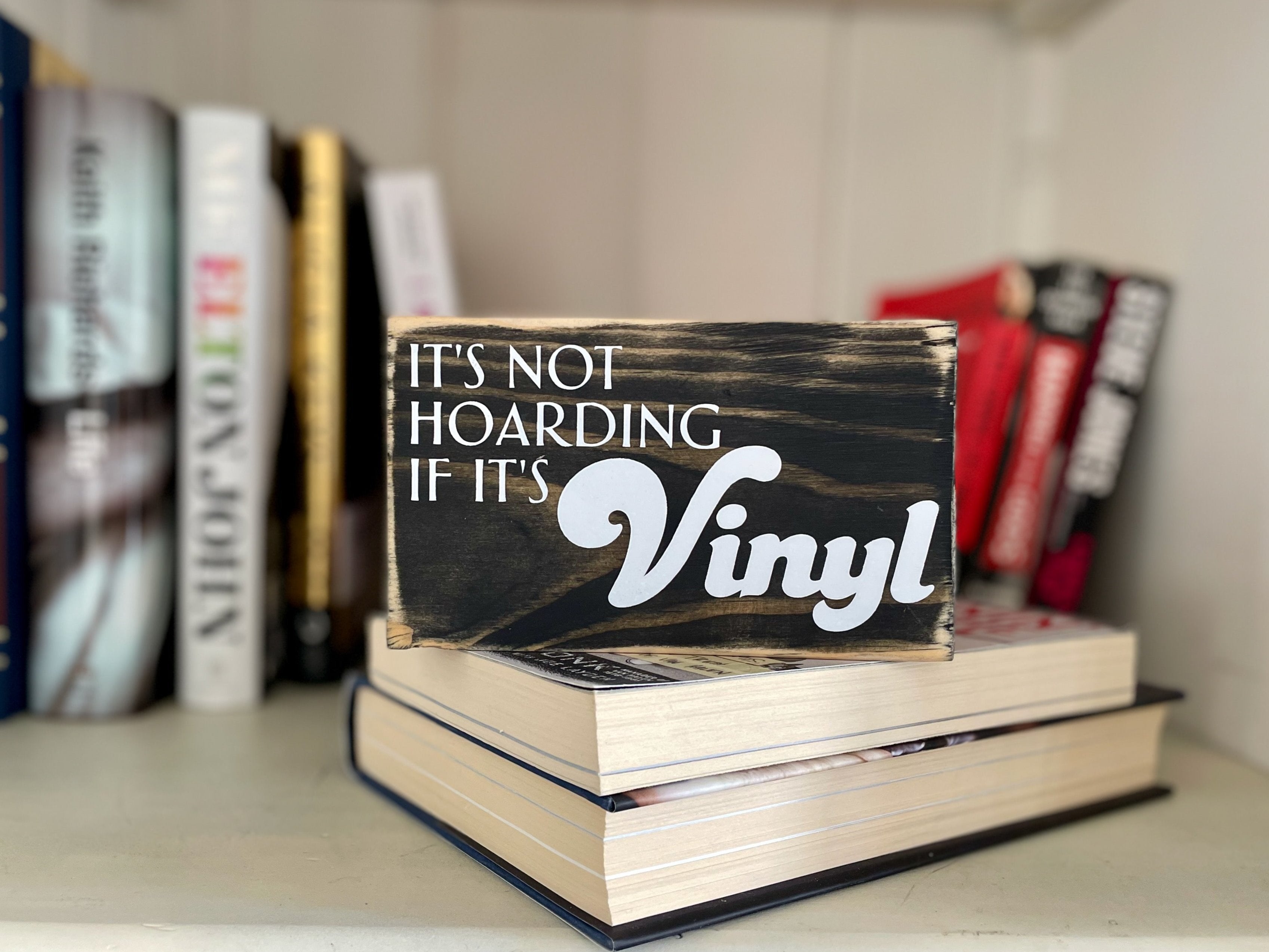 A small, black, wood sign sits on a stack of books; rock star biographies are blurry in the background. In white, the sign reads, "It's not hoarding if it's vinyl". 