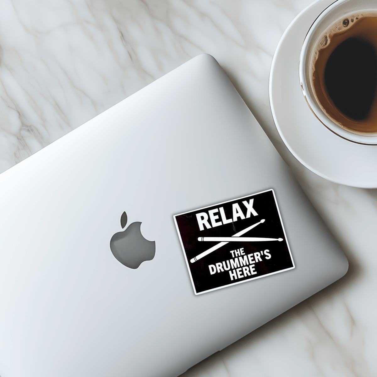 Laptop with 'Relax, The Drummer's Here' sticker next to a cup of coffee on a marble surface