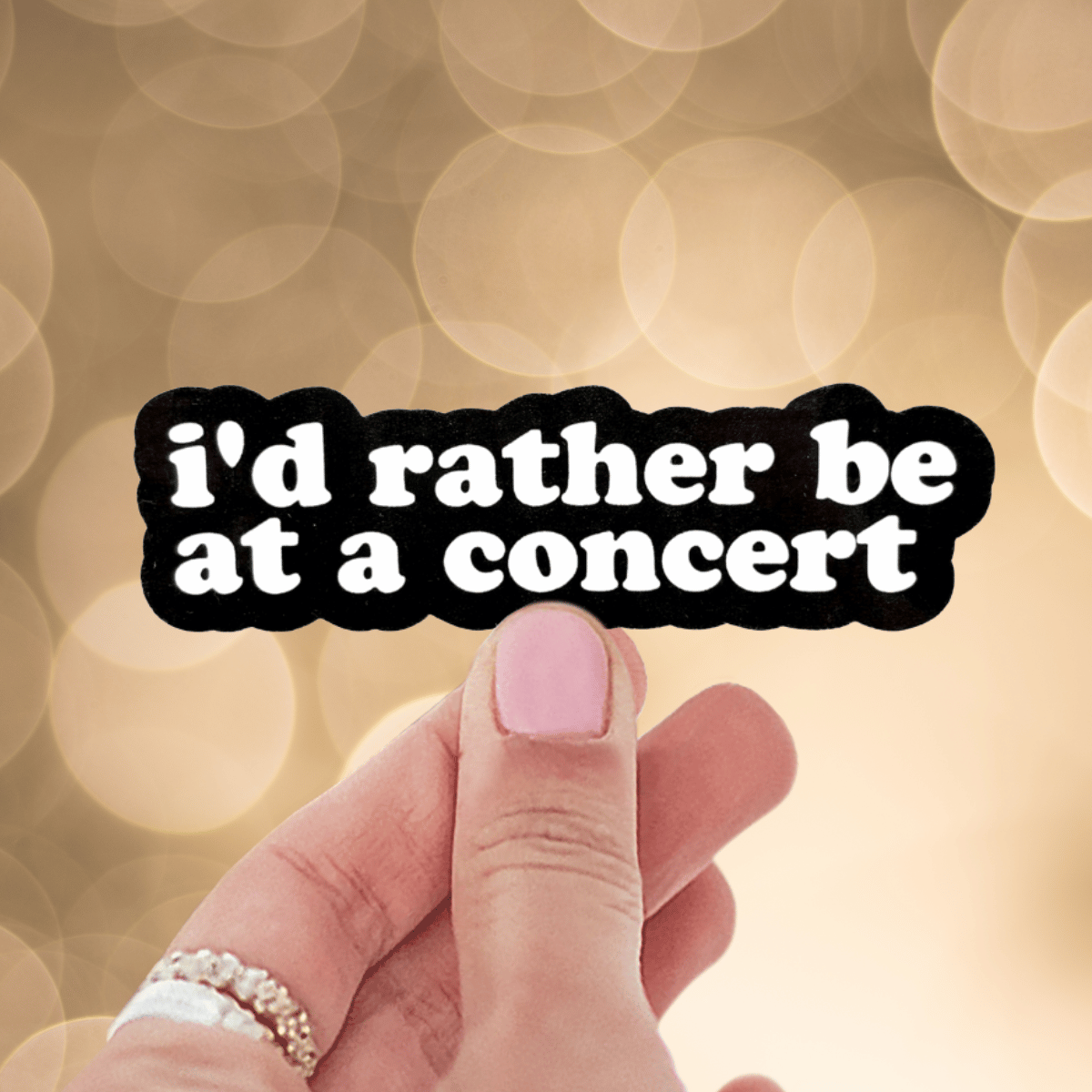 Hand holding a sticker with 'I'd rather be at a concert' text against a blurred background.
