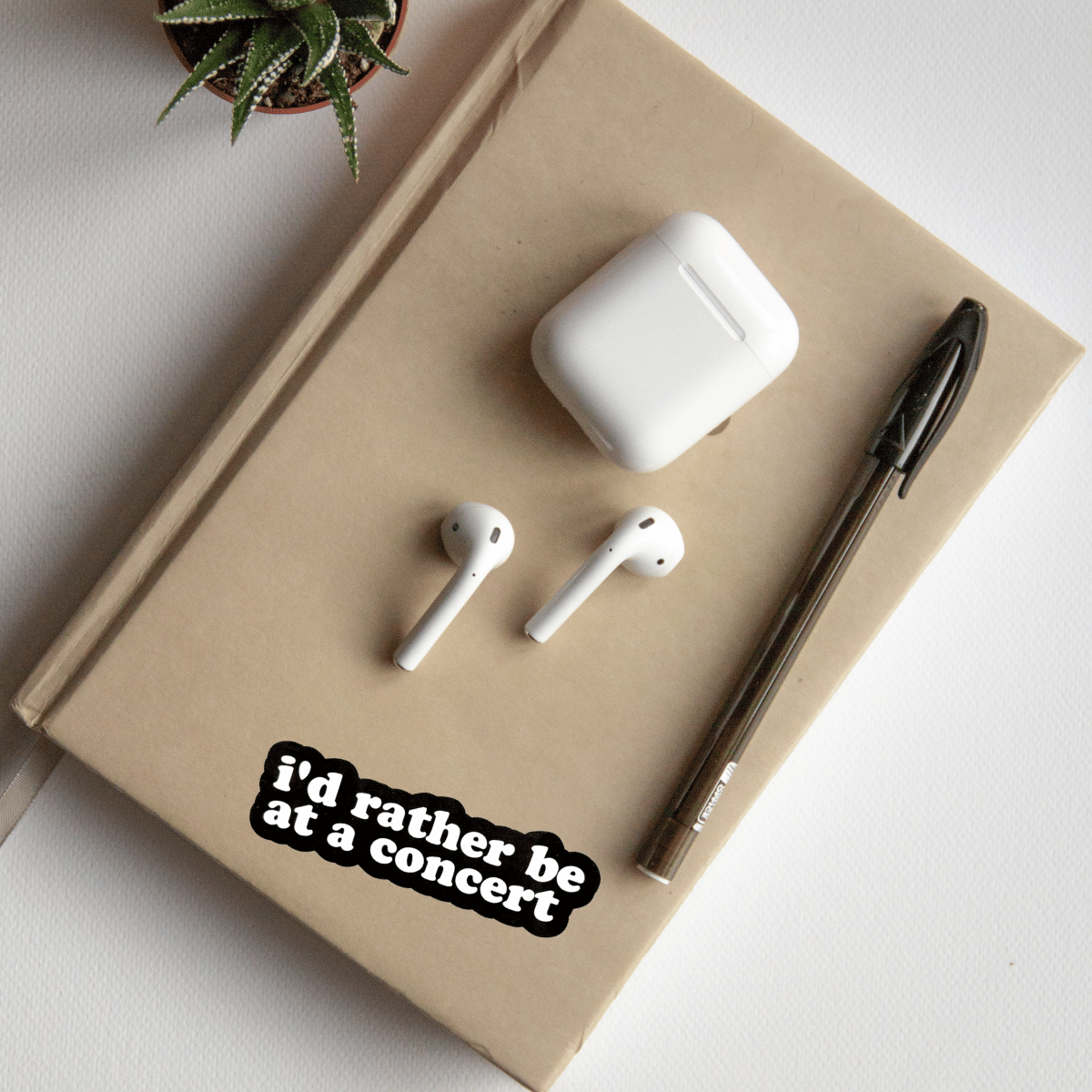 White wireless earbuds and case on a beige notebook with a sticker saying 'i'd rather be at a concert' on a light background.