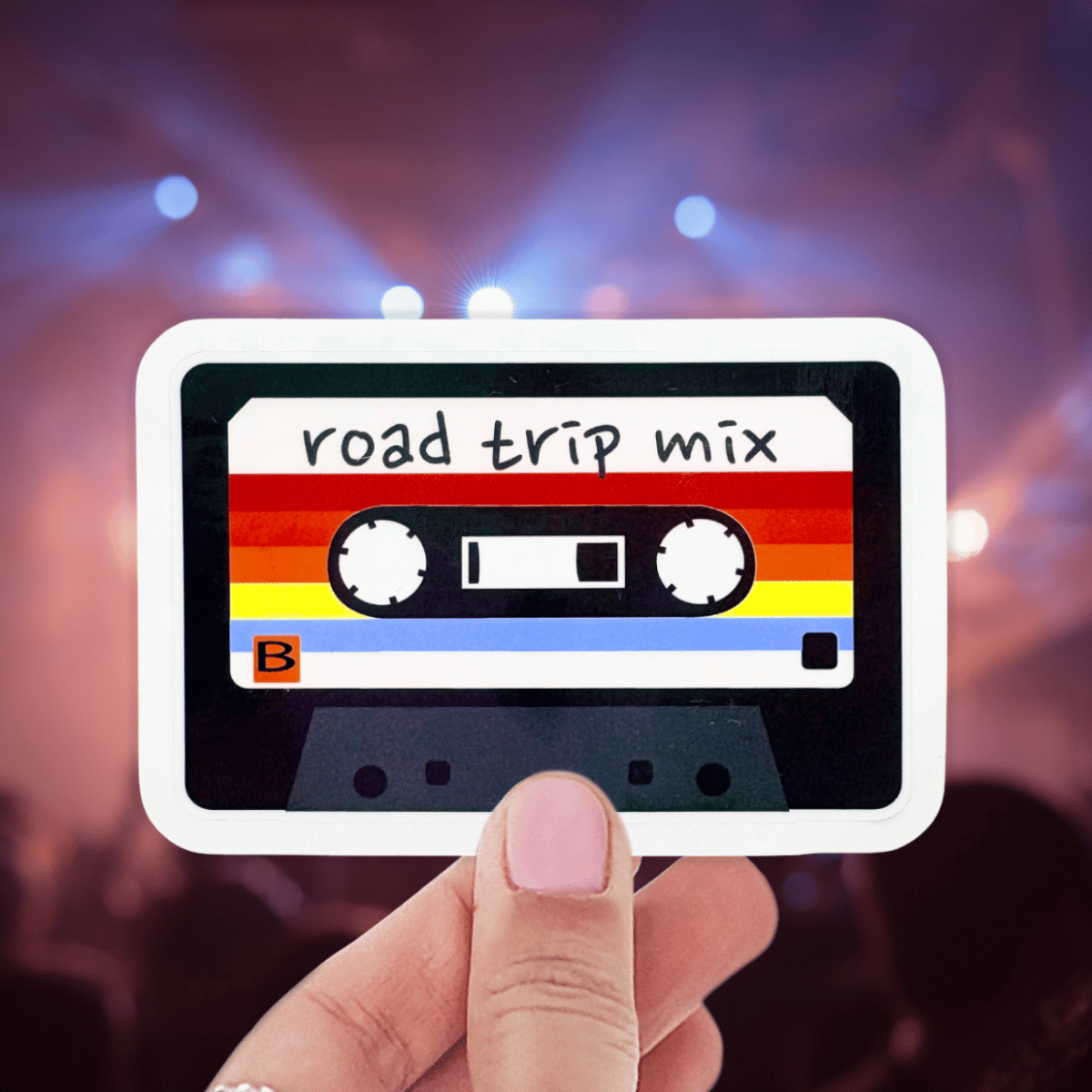 Cassette tape sticker labeled 'road trip mix' held by a hand with blurred concert lights in the background.