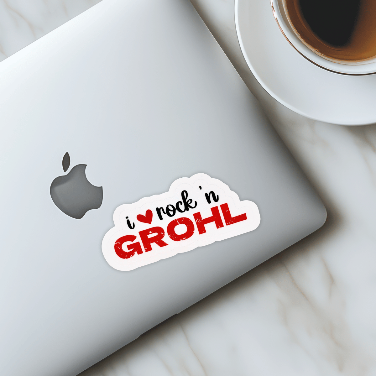The "I love Rock n Grohl" sticker is on a laptop sitting on a marble counter next to a cup of coffee. 