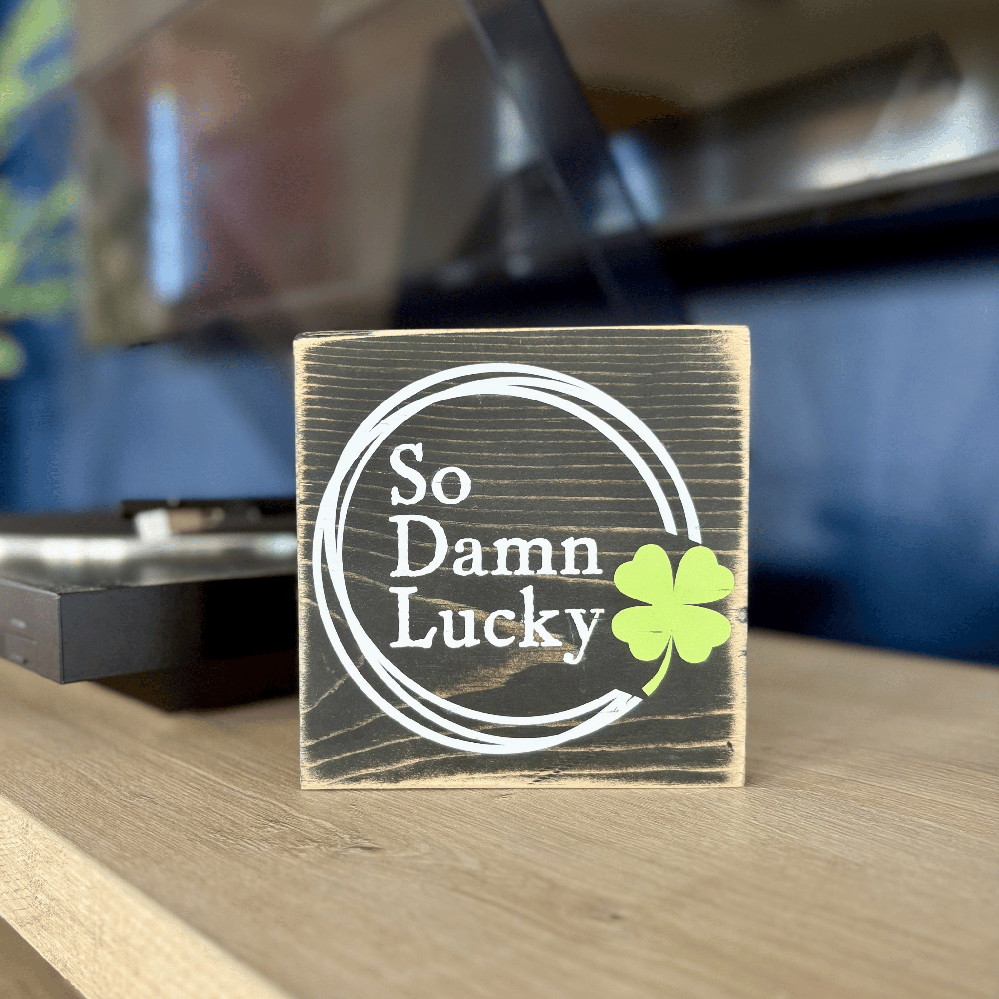 Wooden block with 'So Damn Lucky' and a clover leaf on a table.