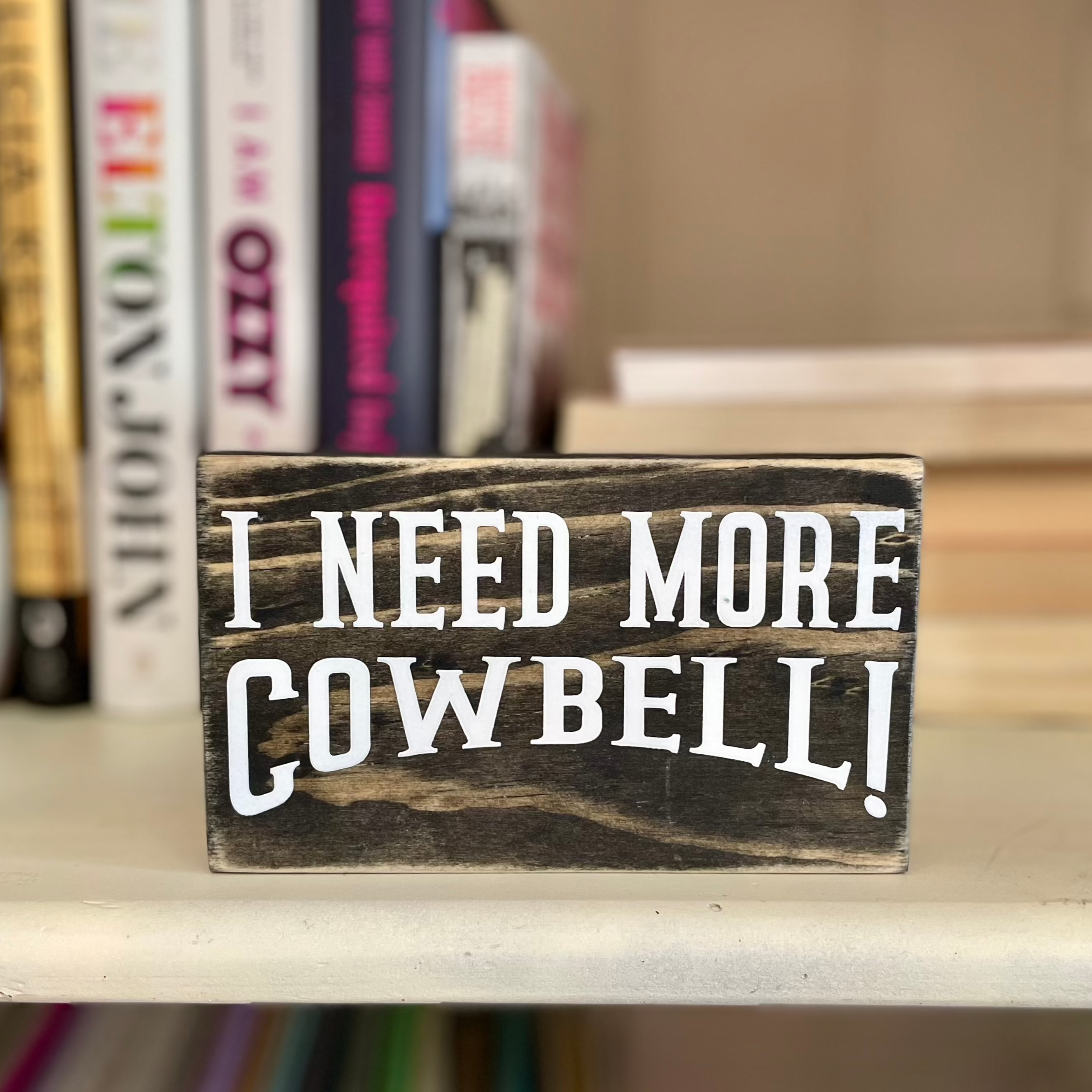 A small, black, wood sign sits on a bookshelf with books blurred in the background. 