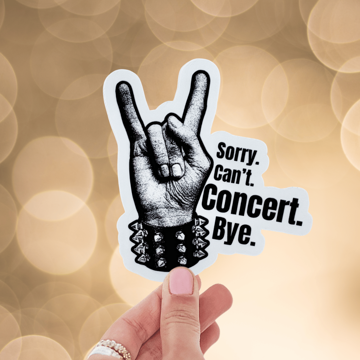 Hand holding a sticker with a rock hand gesture and text 'Sorry. Can't. Concert. Bye.' against a blurred background.