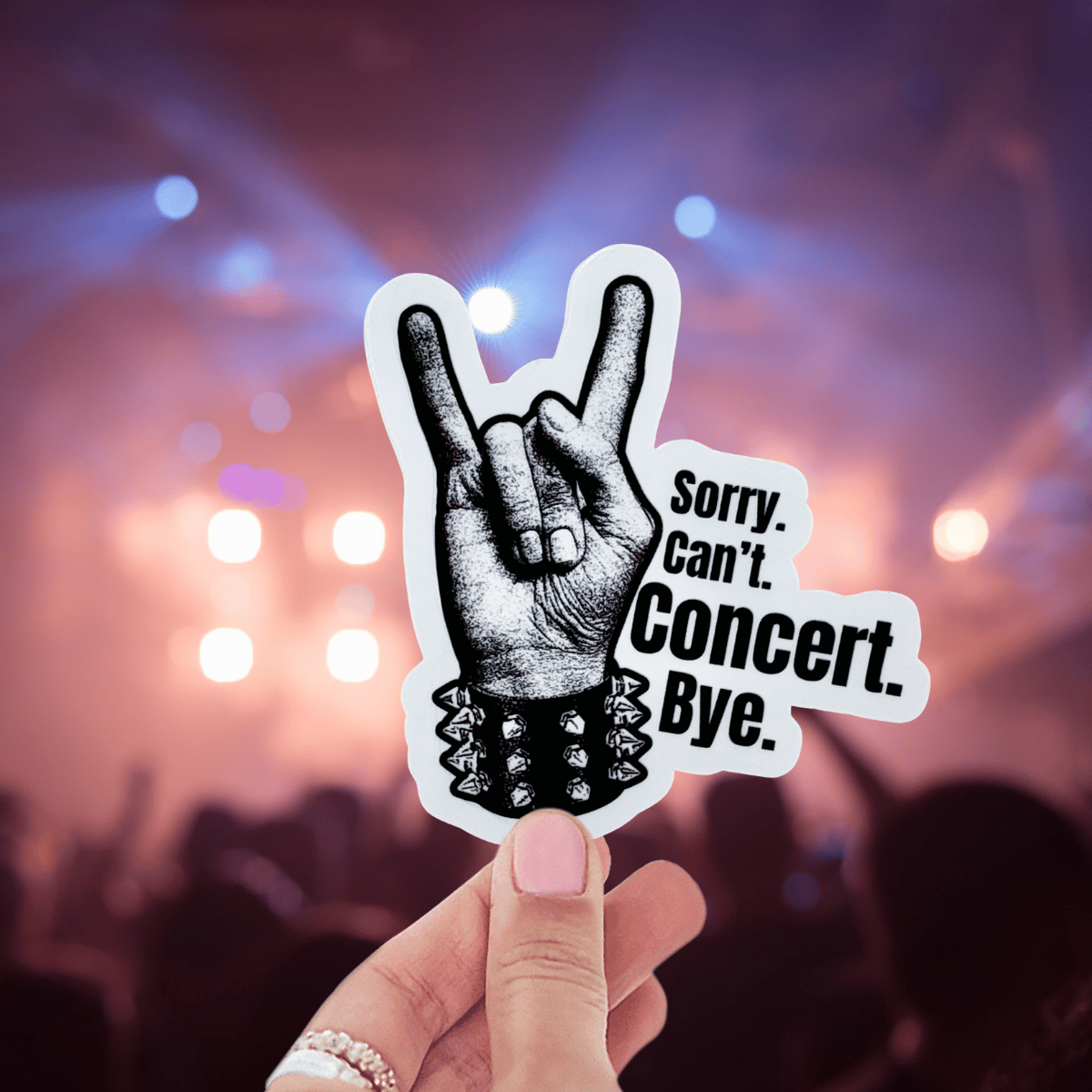 Hand holding a sticker with a rock hand gesture and text 'Sorry. Can't. Concert. Bye.' against a concert background.