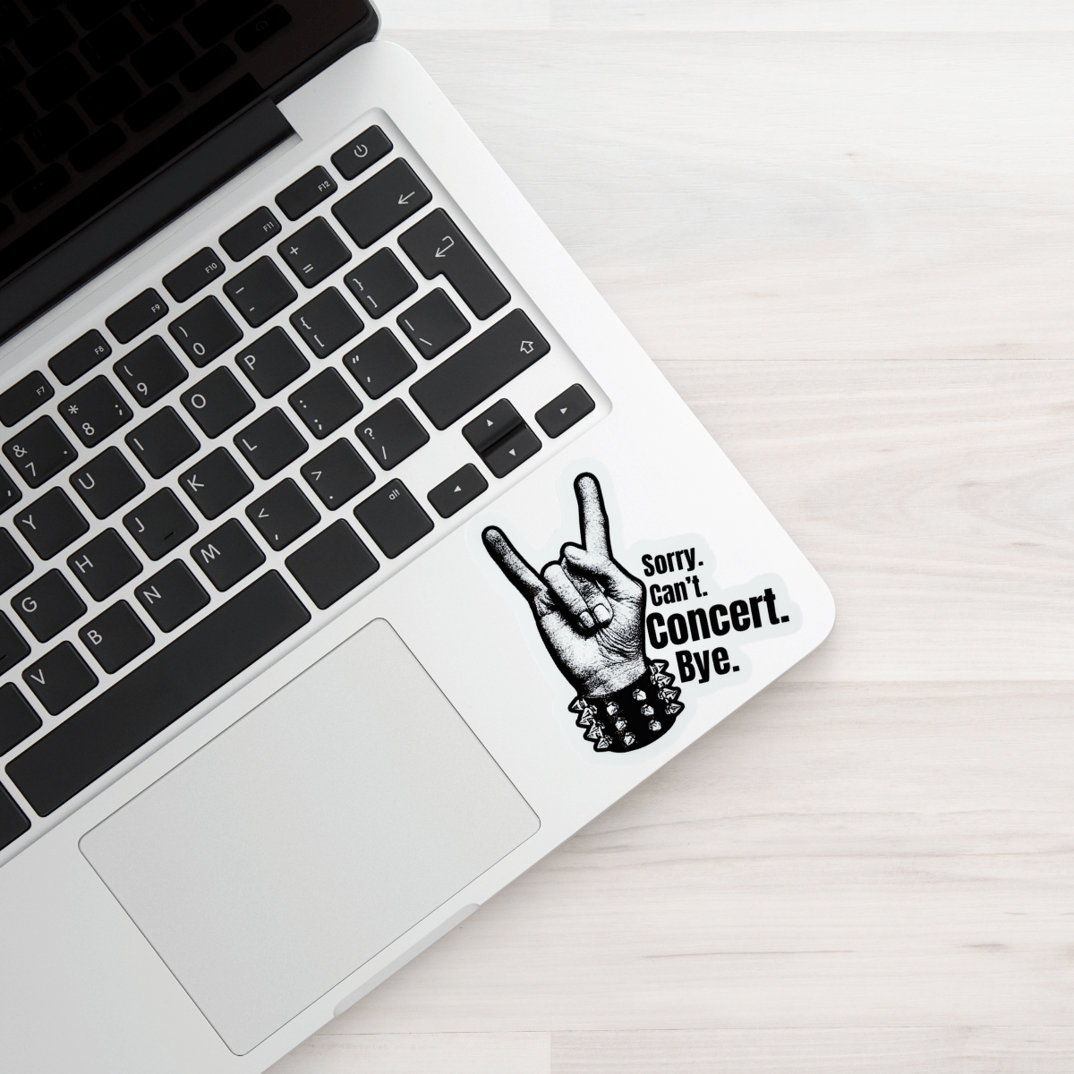 Laptop with a sticker of a hand making the sign of the horns and text 'Sorry, Can't. Concert is Bye.' on a light wooden surface.