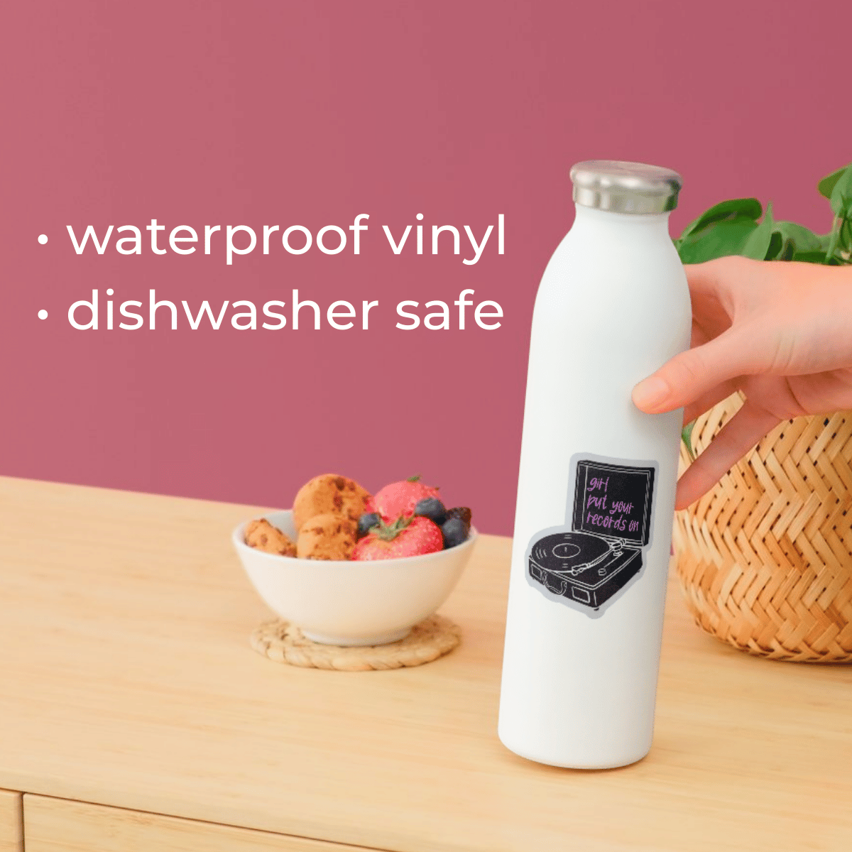 White water bottle with vinyl design held by a hand on a wooden surface with a bowl of fruit and pink background.