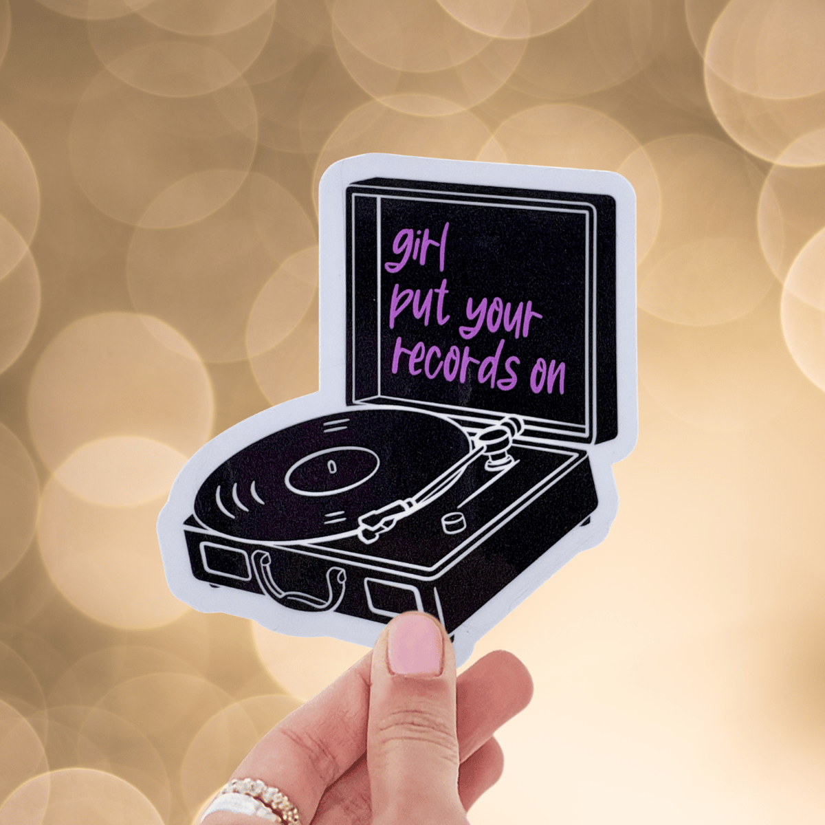 Vinyl record player sticker with 'girl put your records on' text held by a hand against a blurred background.