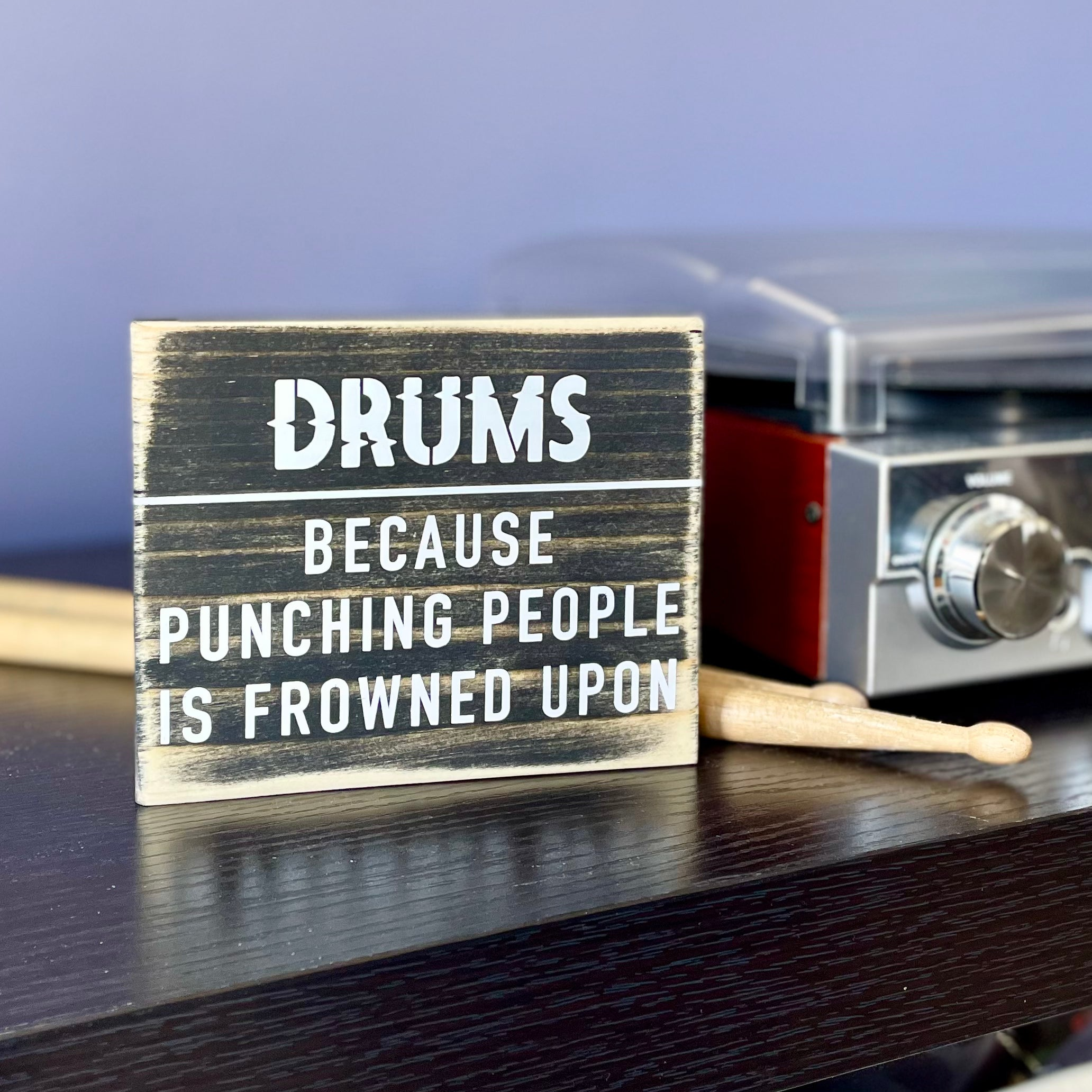 A small, black, wood sign sits on a black table next to a record player and a pair of wood drumsticks. In white it reads, "Drums. Because punching people is frowned upon."