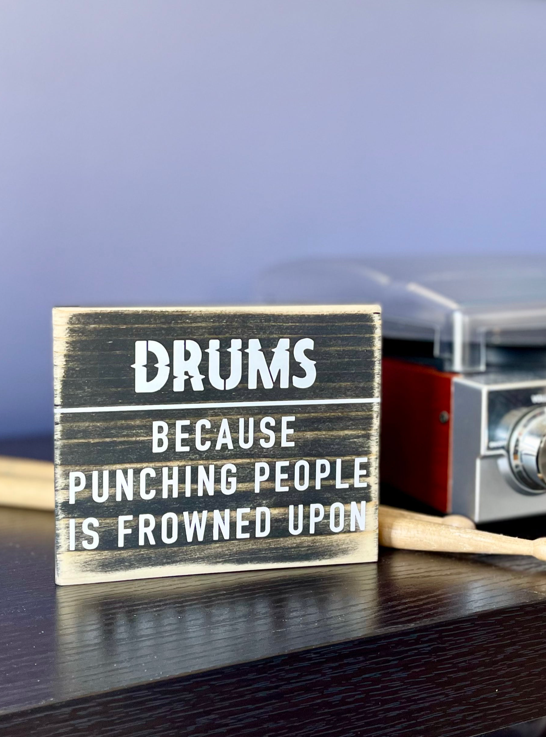 A small, black, wood sign sits on a black table next to a record player and a pair of wood drumsticks. In white it reads, "Drums. Because punching people is frowned upon."