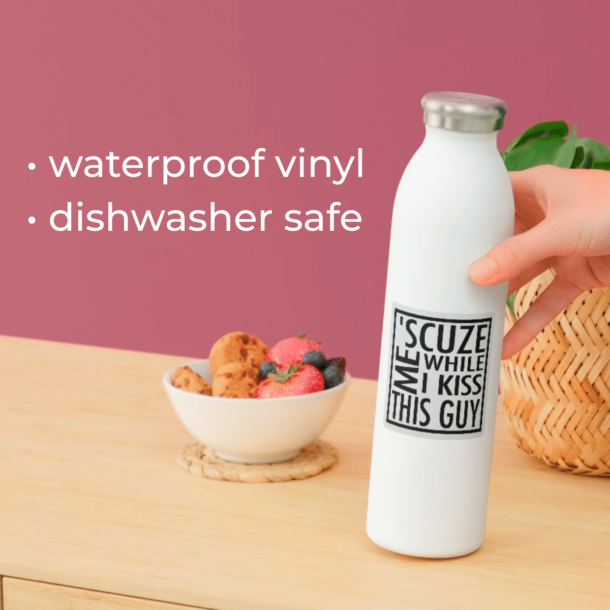 White water bottle with text held by a hand on a wooden surface with a bowl of fruit in the background.